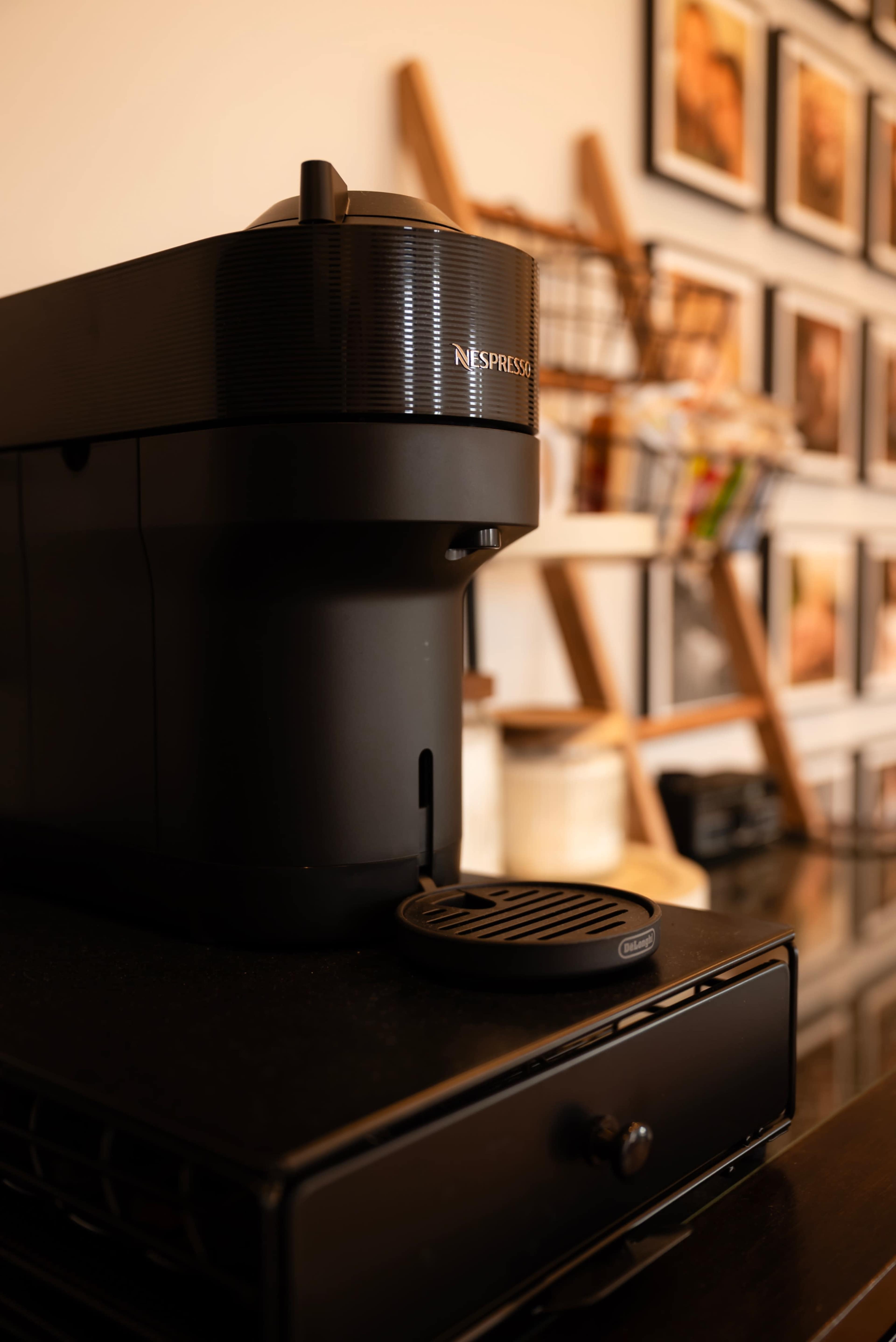 A black Nespresso coffee machine sits on a countertop in front of a wall decorated with framed photographs.