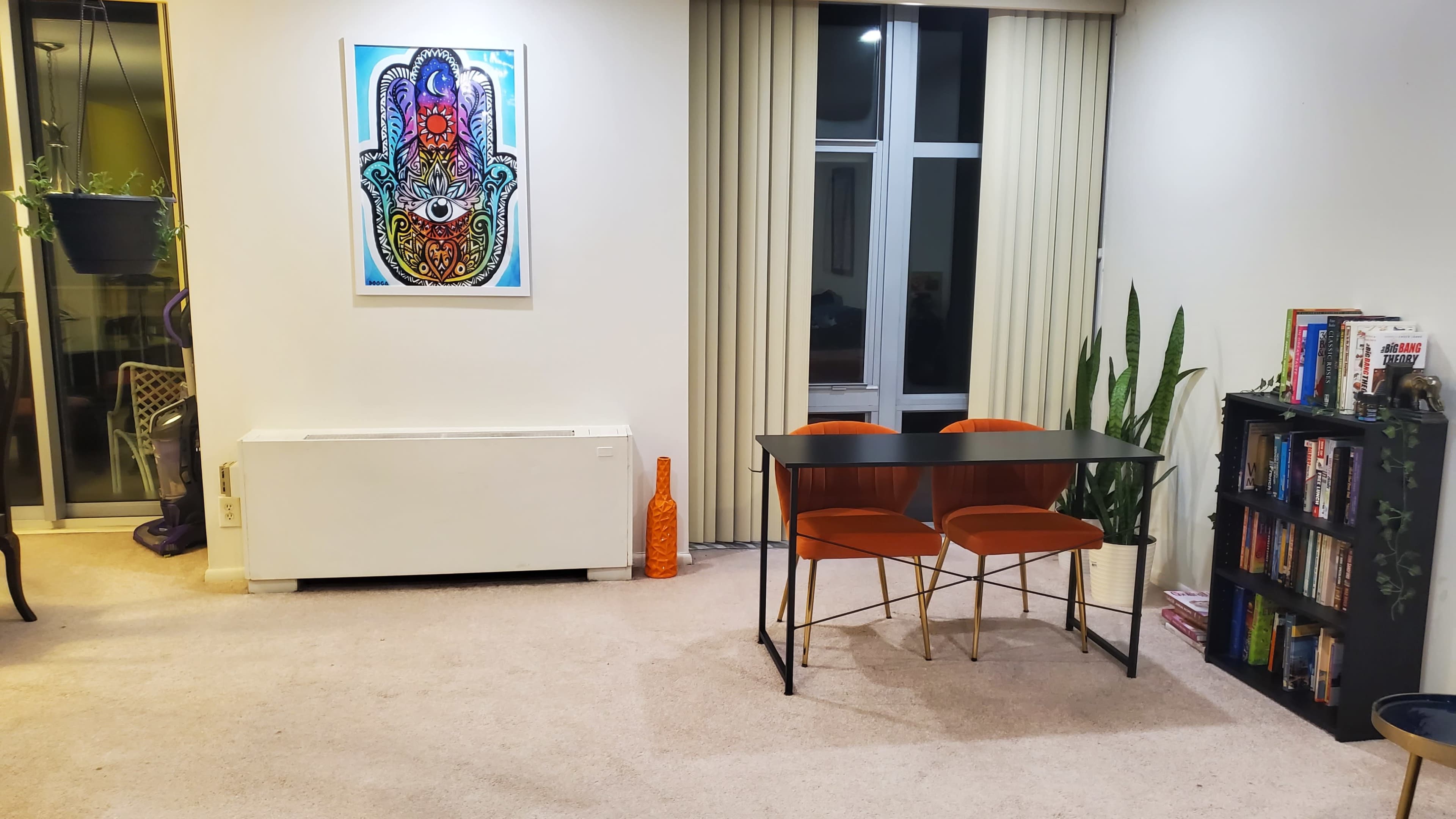 The image shows a minimalist living space with a small dining table and two orange chairs, a potted plant, and a shelf filled with books, against a backdrop of beige walls and vertical blinds.