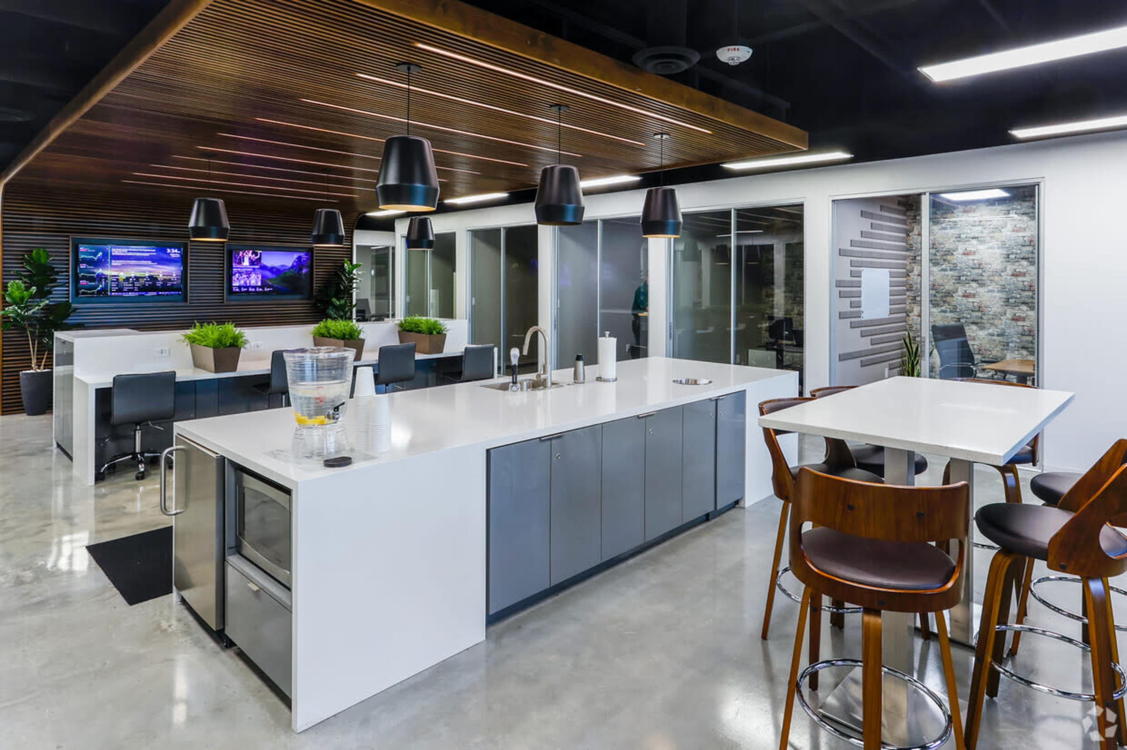 A modern office kitchen area features a white countertop, gray cabinetry, and seating with wooden chairs, alongside two screens displaying images.