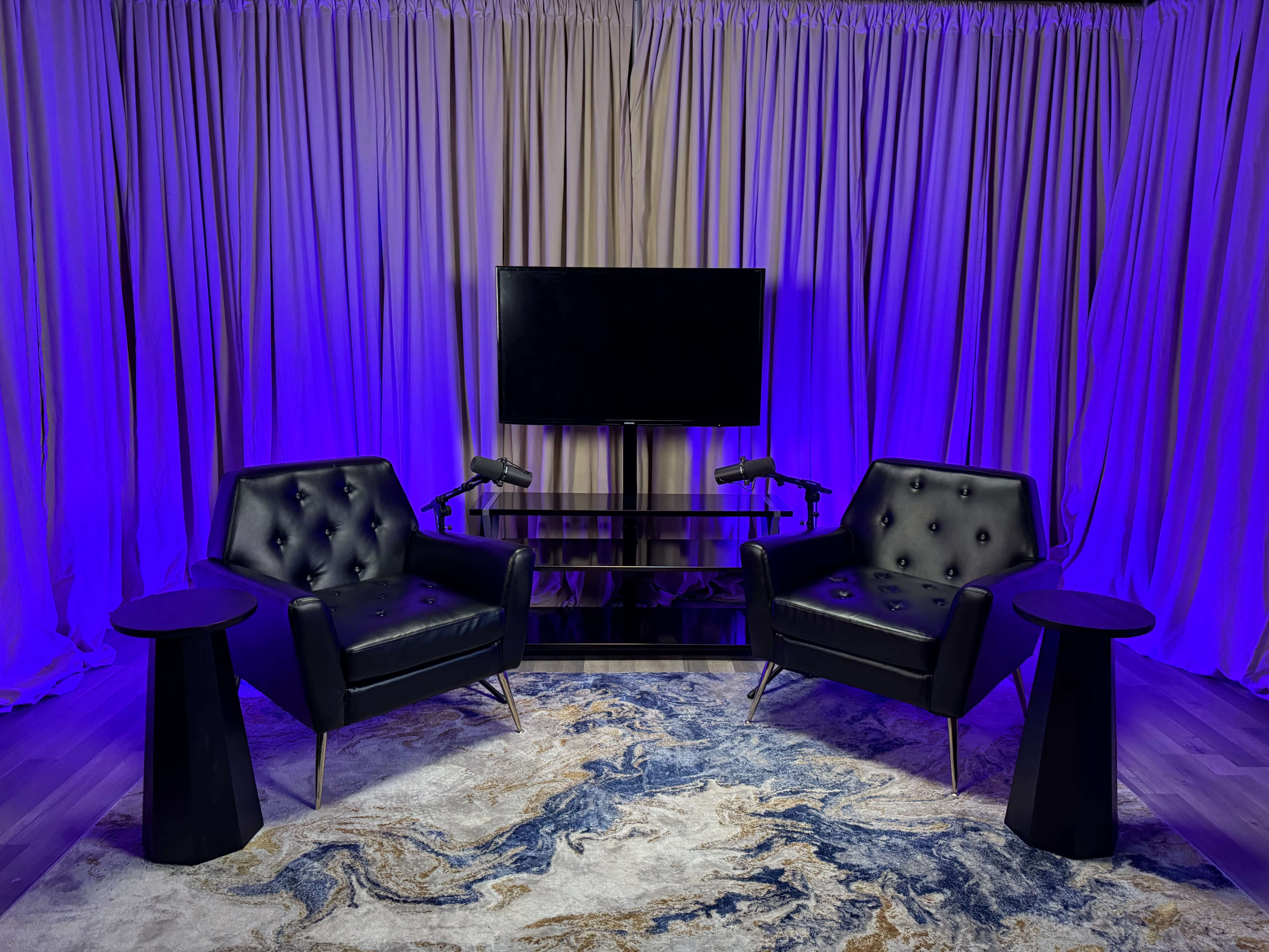 The image shows a modern interview setup featuring two black tufted armchairs, two small tables, and a television, all set against a backdrop of purple lighting and draped fabric.