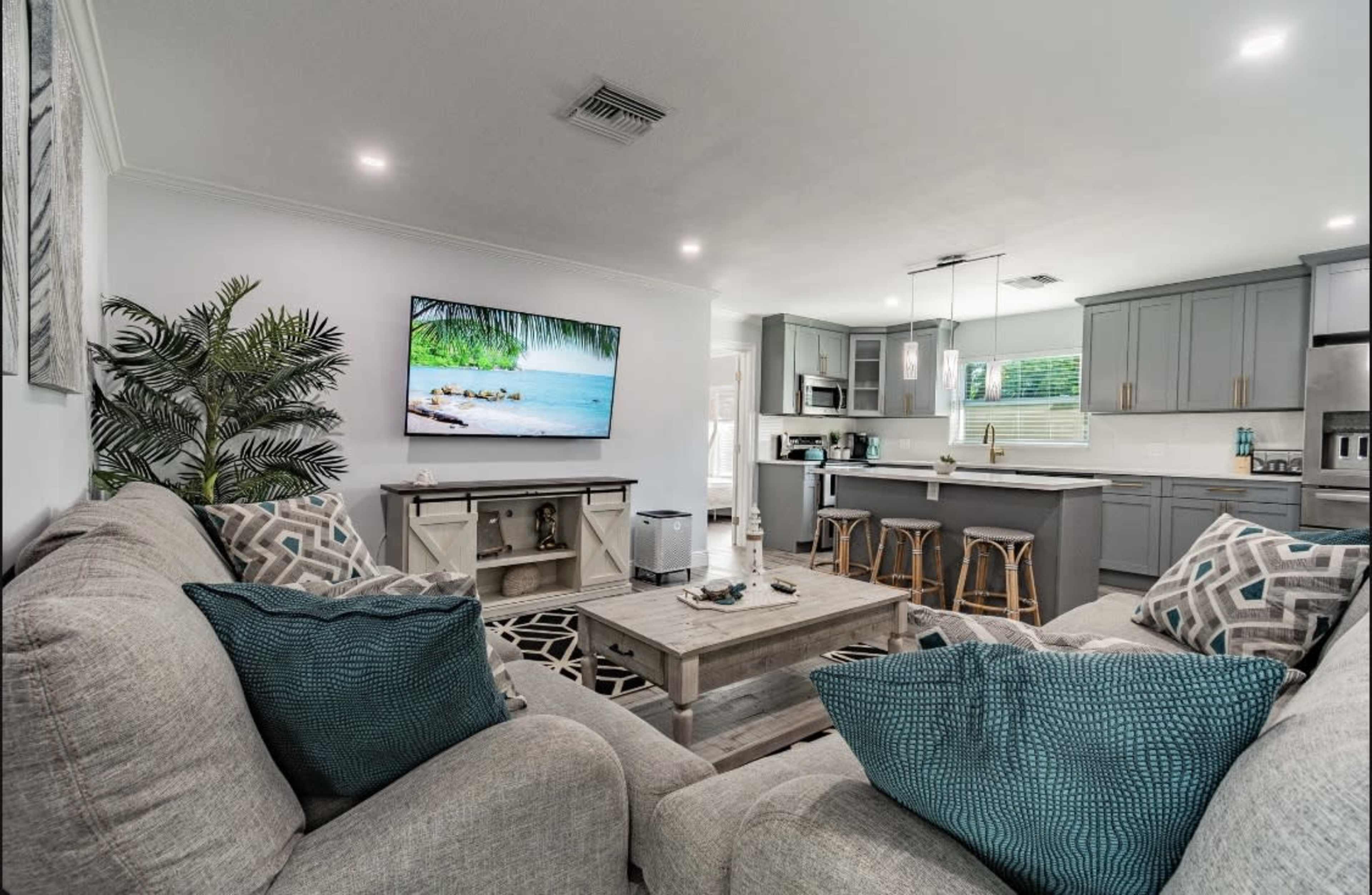 The image shows a modern living room with a large TV mounted on the wall, a gray sectional sofa with decorative pillows, a coffee table, and an open kitchen area in the background.