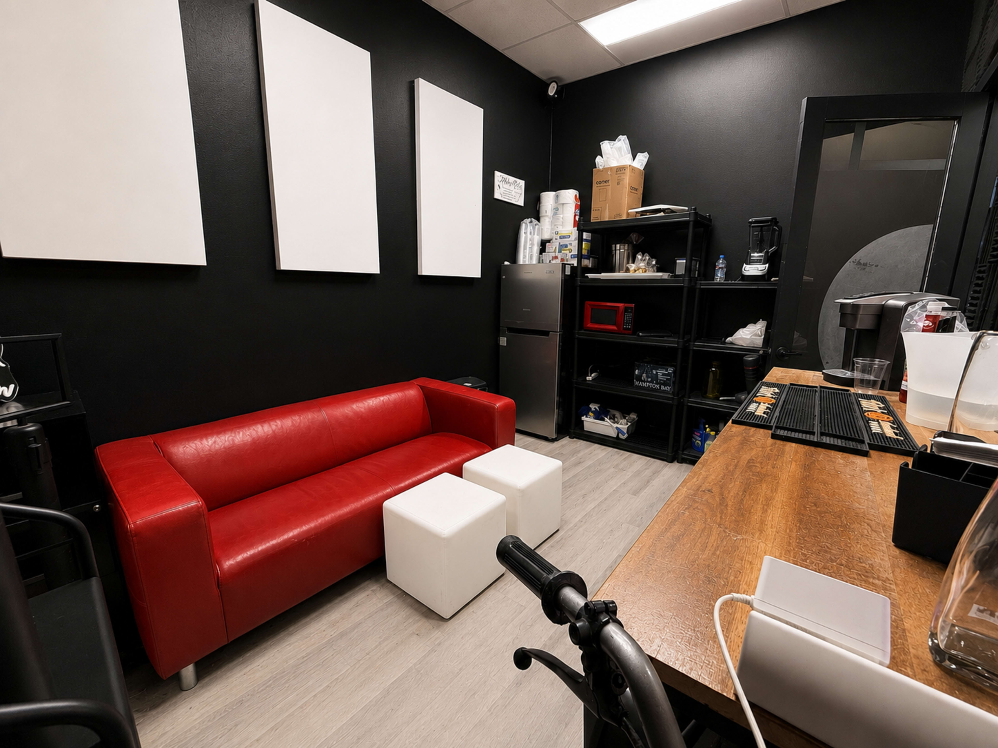 The image shows a small room with a red sofa, two white stools, and a workspace featuring a wooden table and shelves with various items.