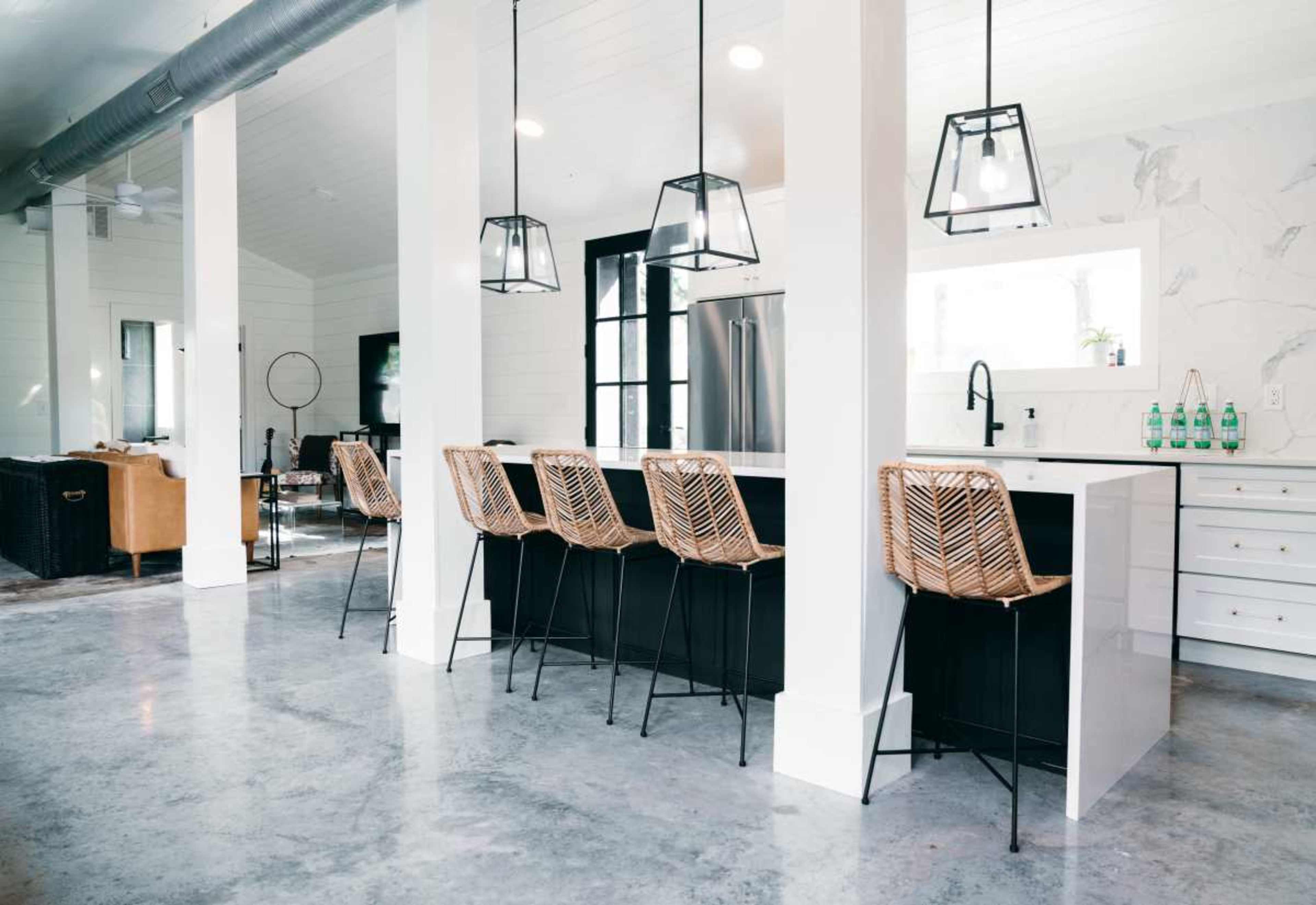 A modern kitchen features a minimalist design with a central island, bar stools, and pendant lighting, complemented by a spacious layout and polished concrete flooring.