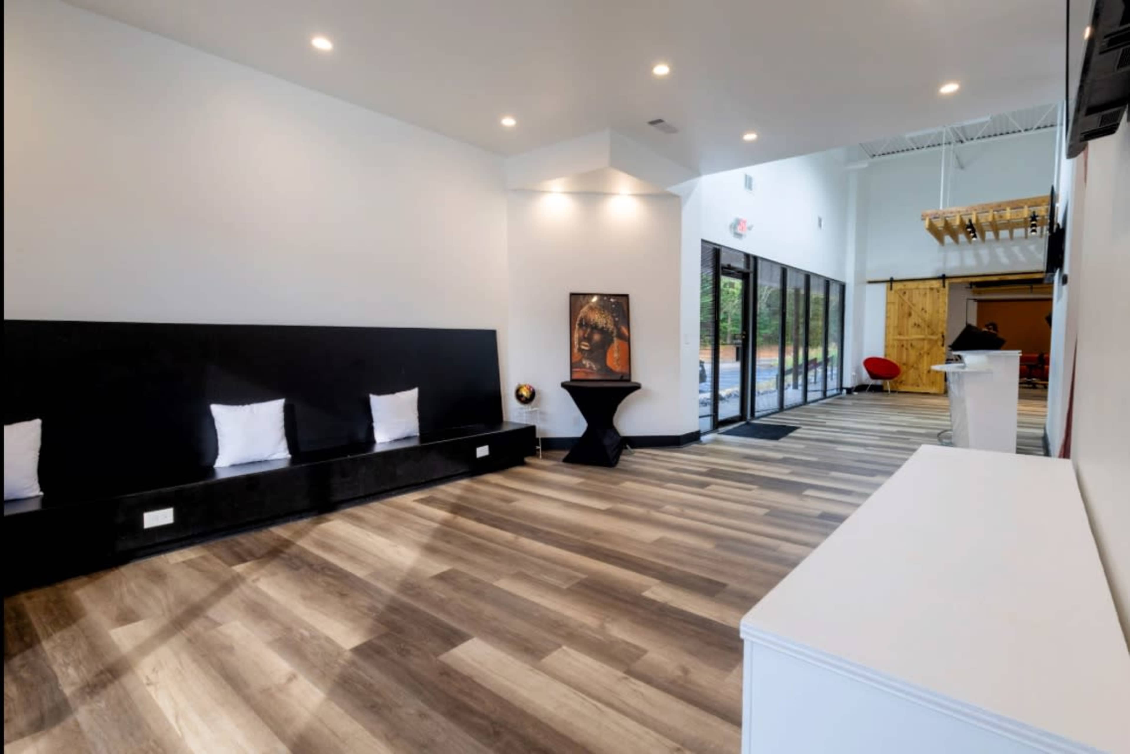 The image shows a modern interior space featuring a wooden floor, black seating along one wall, and large glass doors leading to an outdoor area.