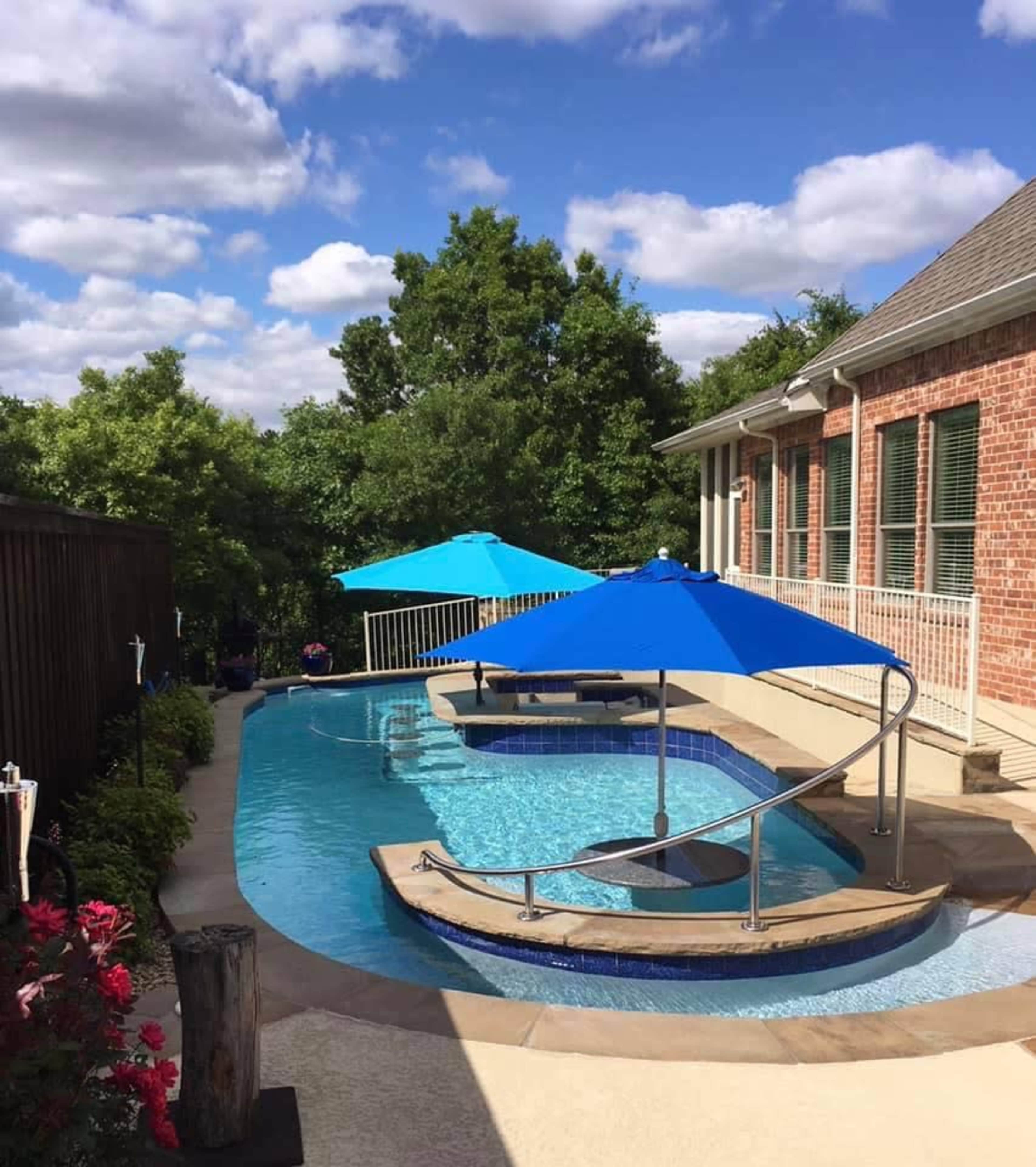 A curved pool with a swim-up bar features two blue umbrellas and is surrounded by greenery and a brick house.