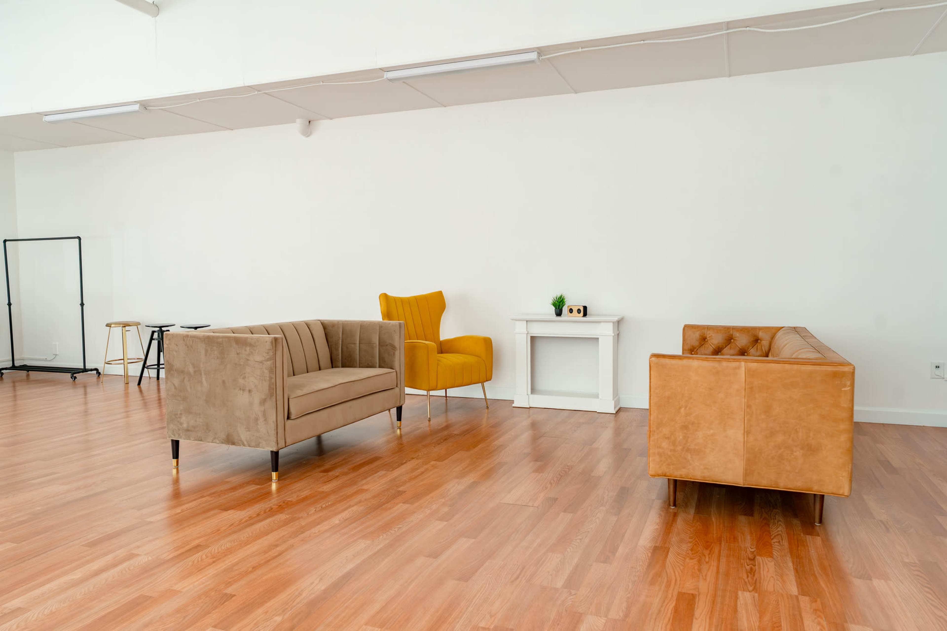 The scene features three distinct pieces of seating furniture: a beige couch, a yellow armchair, and a tan leather couch, arranged in a spacious room with wooden flooring and minimal decor.