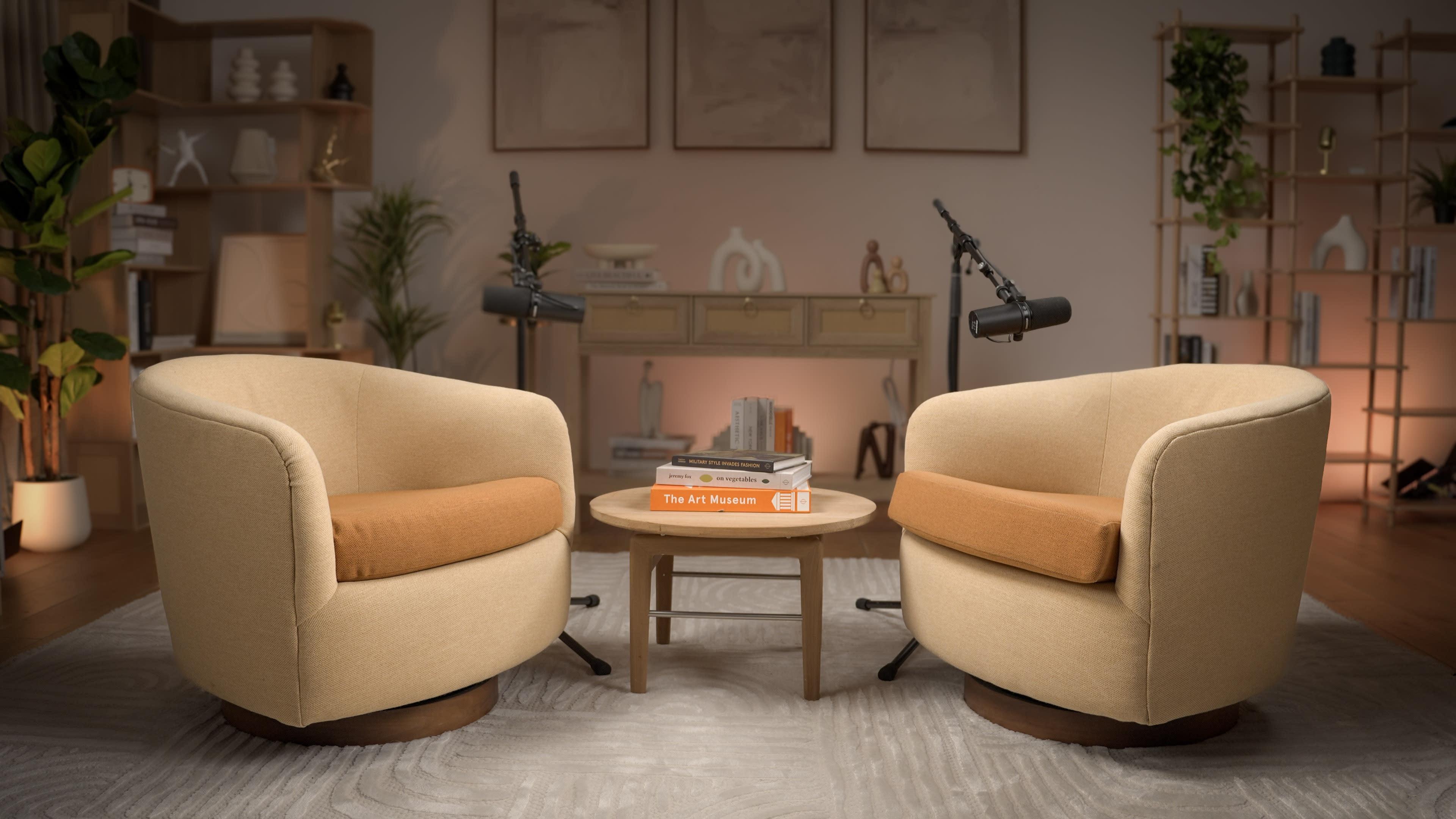 The image shows two beige armchairs with orange cushions facing a small round table in a warmly lit room with a shelf and decorative items in the background.