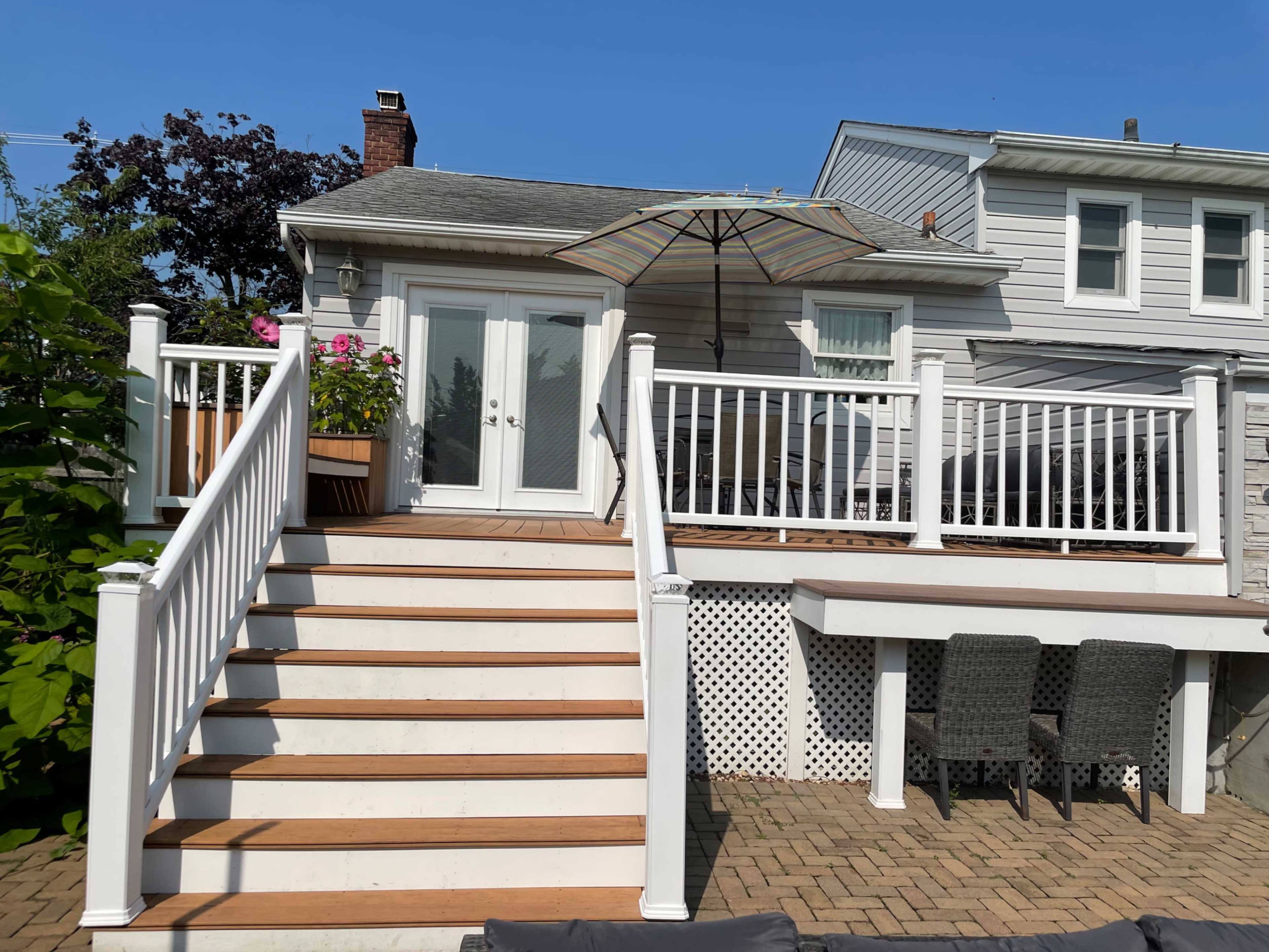 A set of wooden stairs leads up to a deck with white railings and an umbrella, adjacent to a house with a stone and siding exterior.