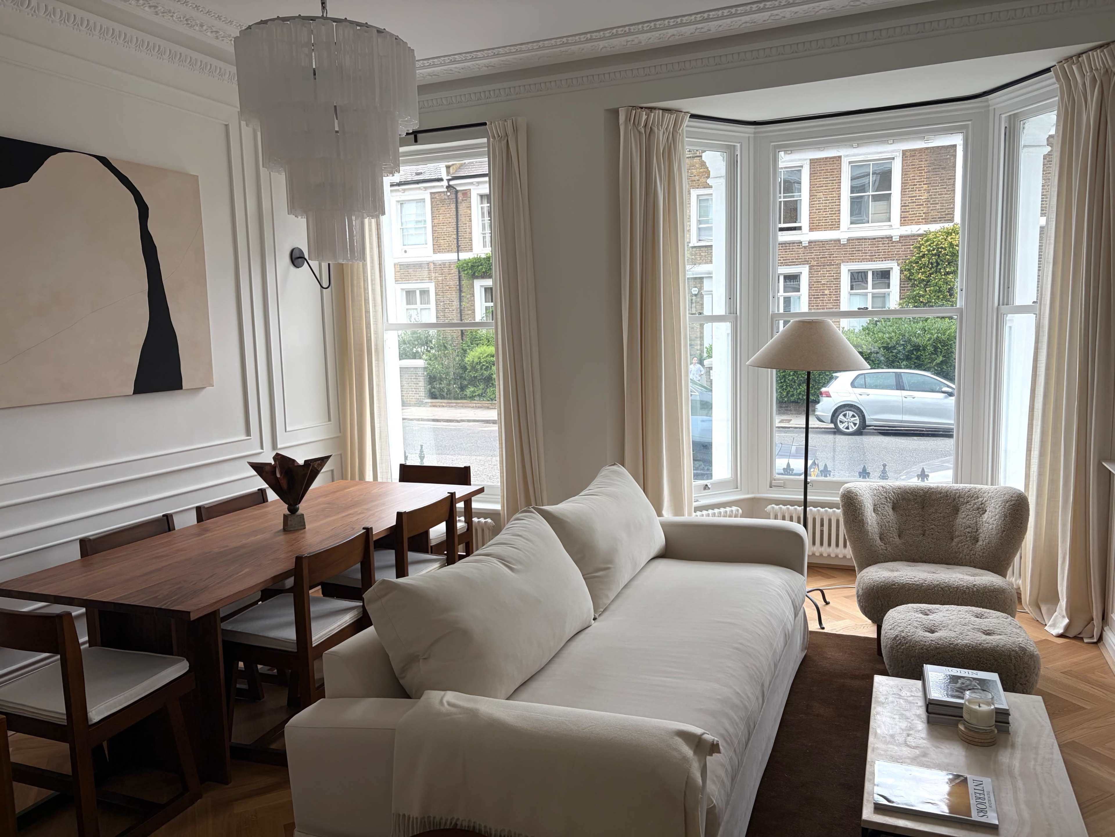 The image shows a modern living room with a white sofa, a wooden dining table, and large bay windows overlooking a street.