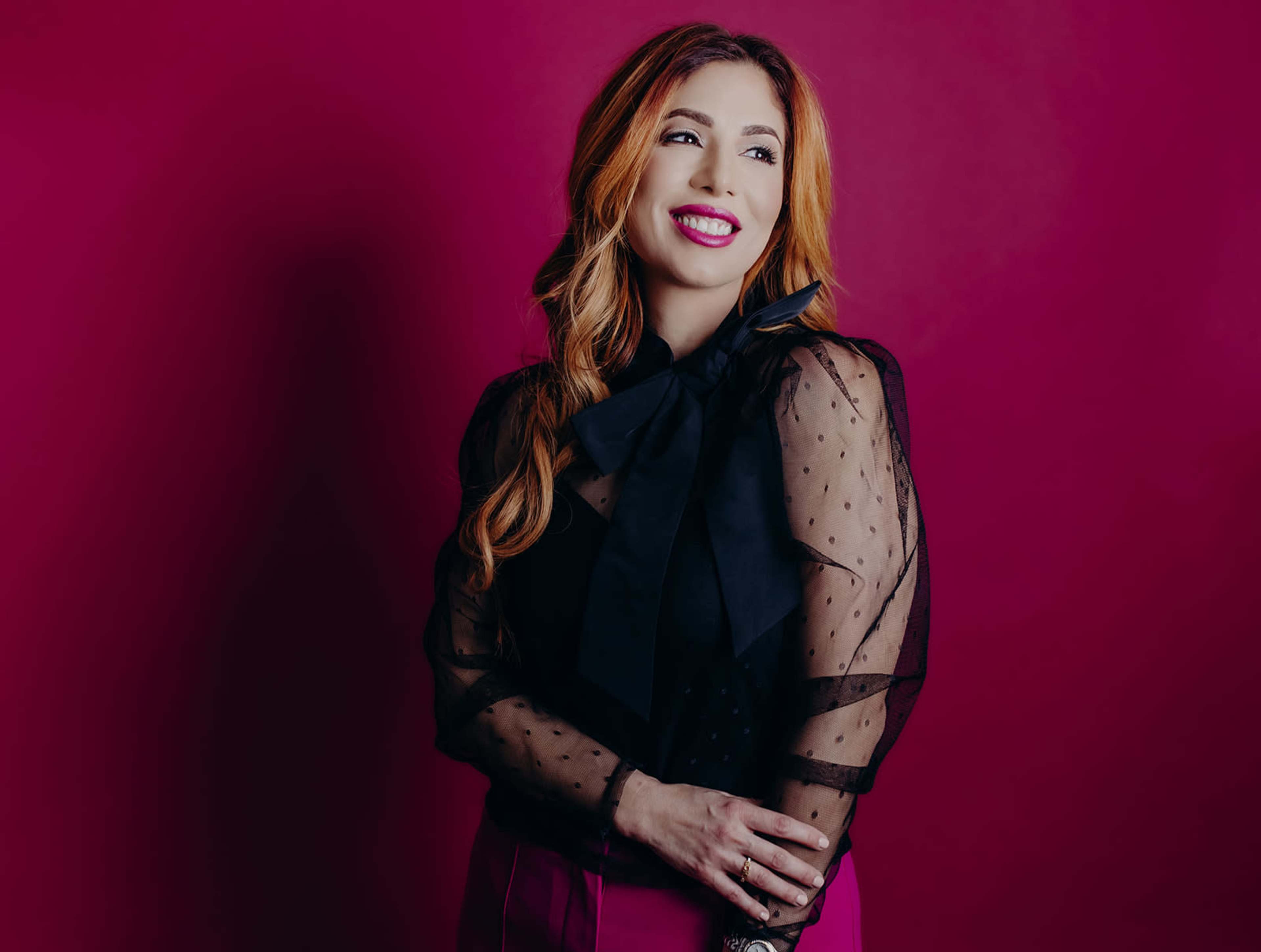 A woman with long, wavy hair smiles while standing against a solid magenta background, wearing a black top with a large bow and transparent sleeves.