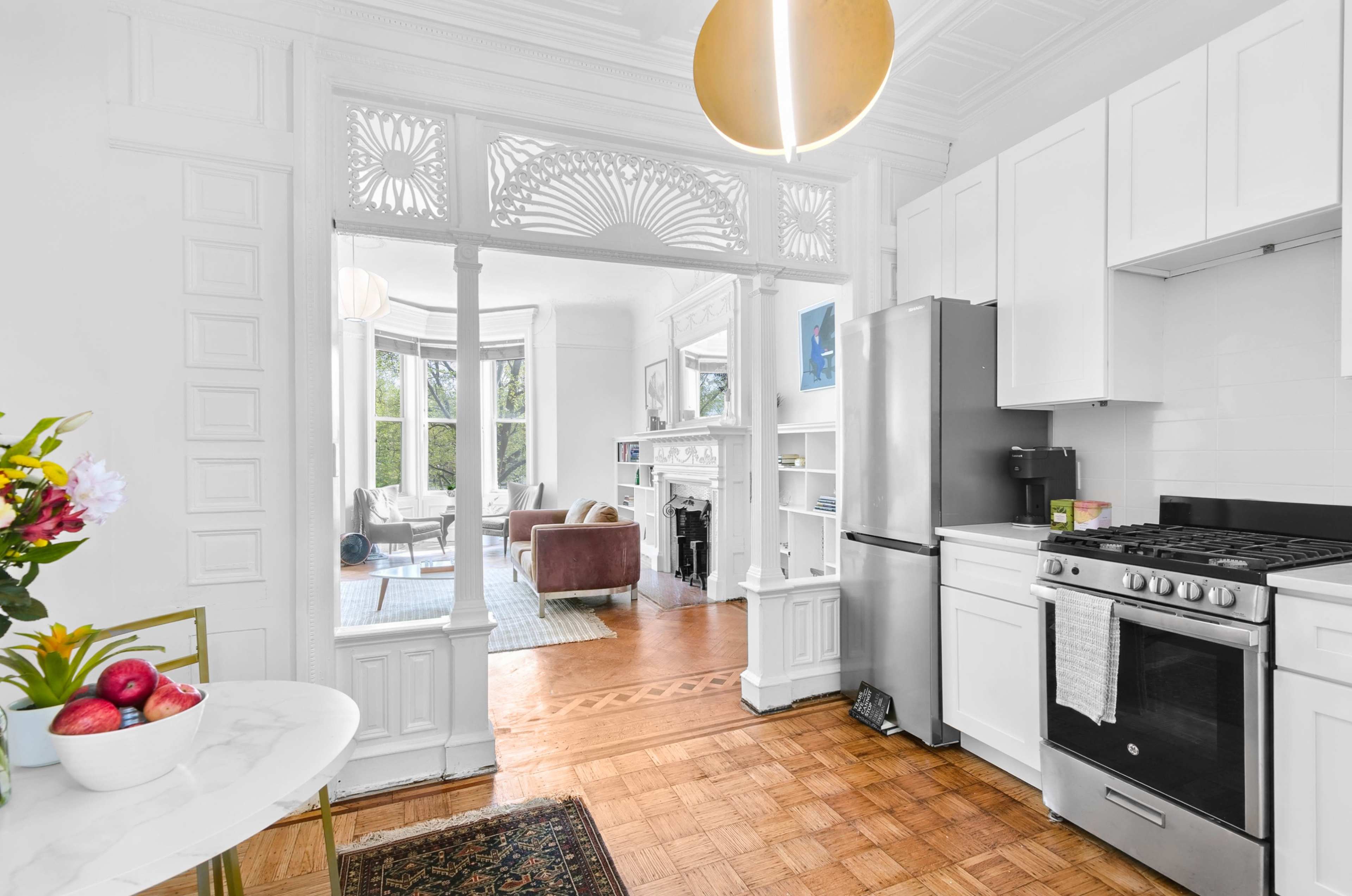 The image shows a bright, modern kitchen connected to a living room through an ornate archway, featuring light-colored cabinetry and hardwood flooring.