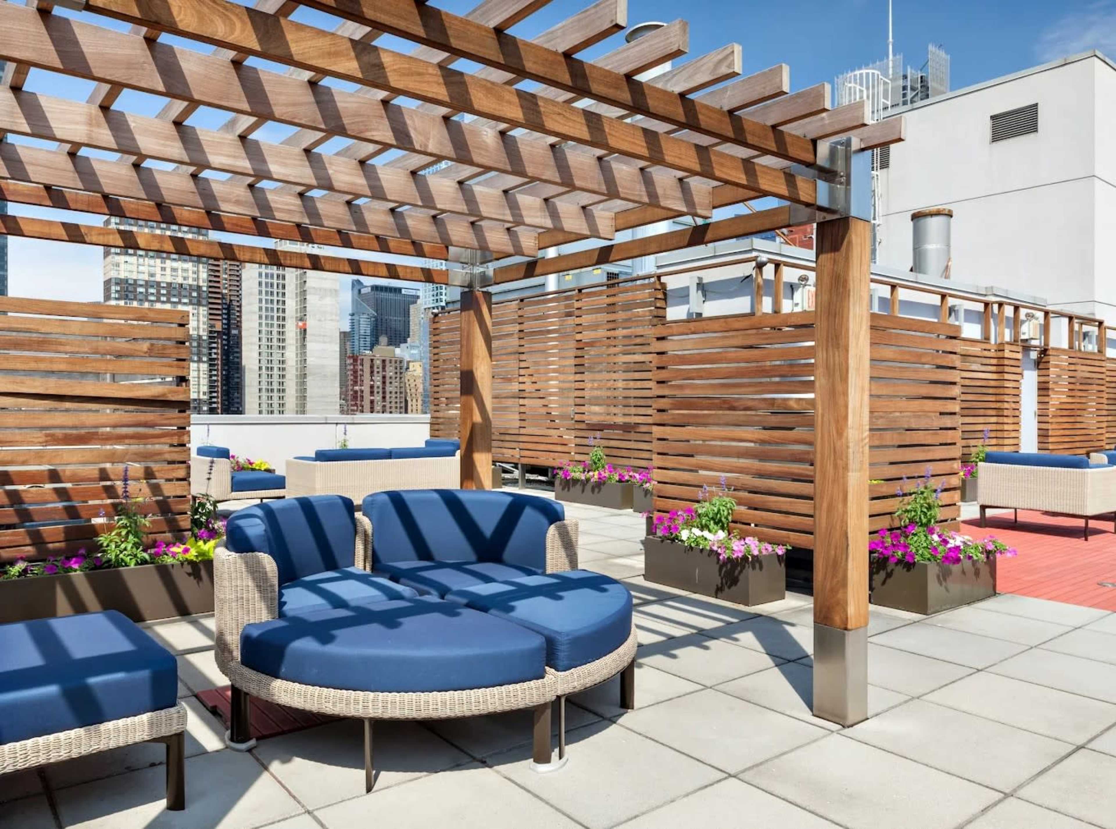 The image shows a rooftop terrace featuring wooden pergolas, circular seating with blue cushions, and planters with colorful flowers against a backdrop of skyscrapers.