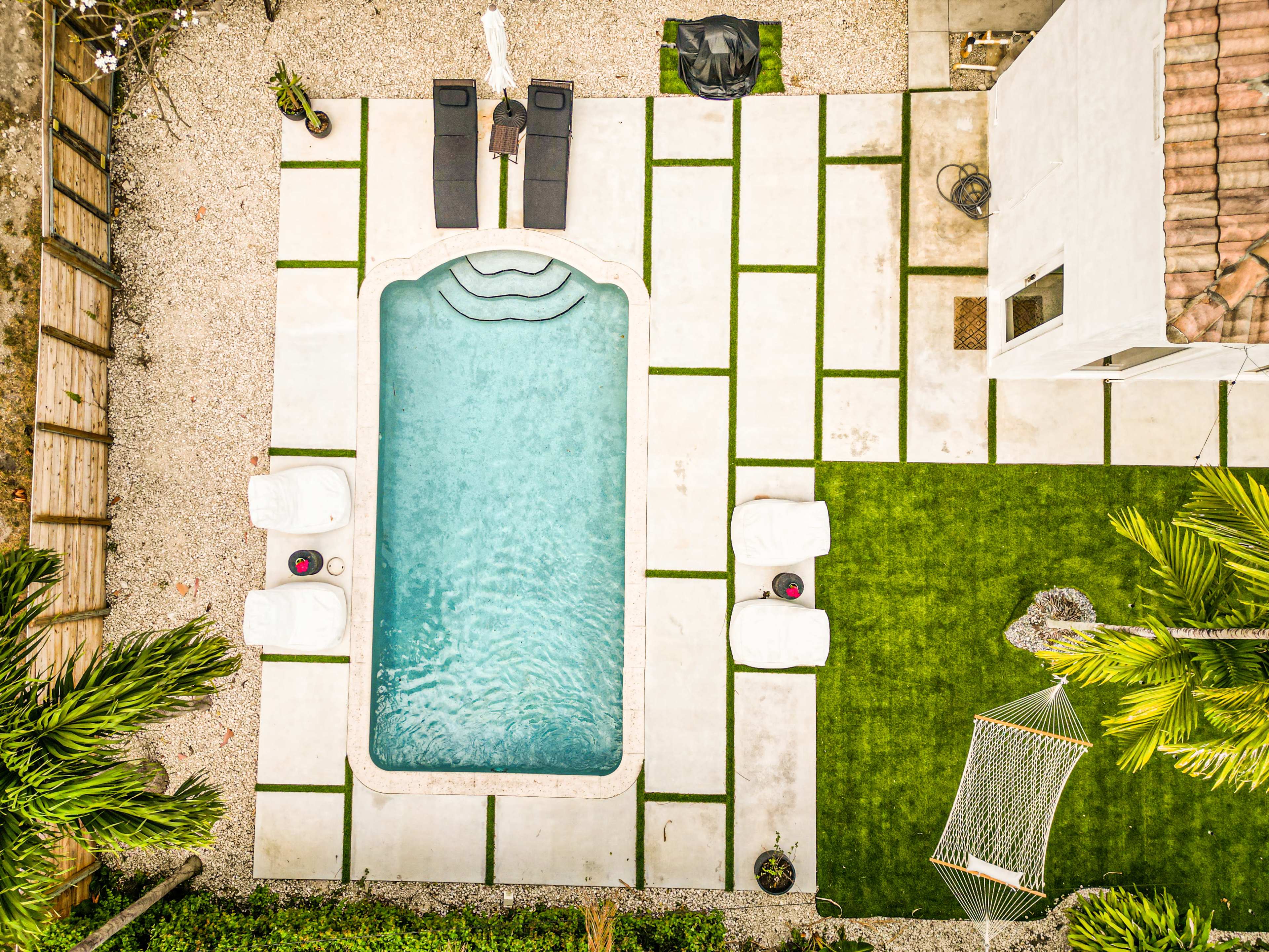 The image shows an aerial view of a rectangular swimming pool surrounded by a stone patio, lounge chairs, and a green lawn with a hammock.