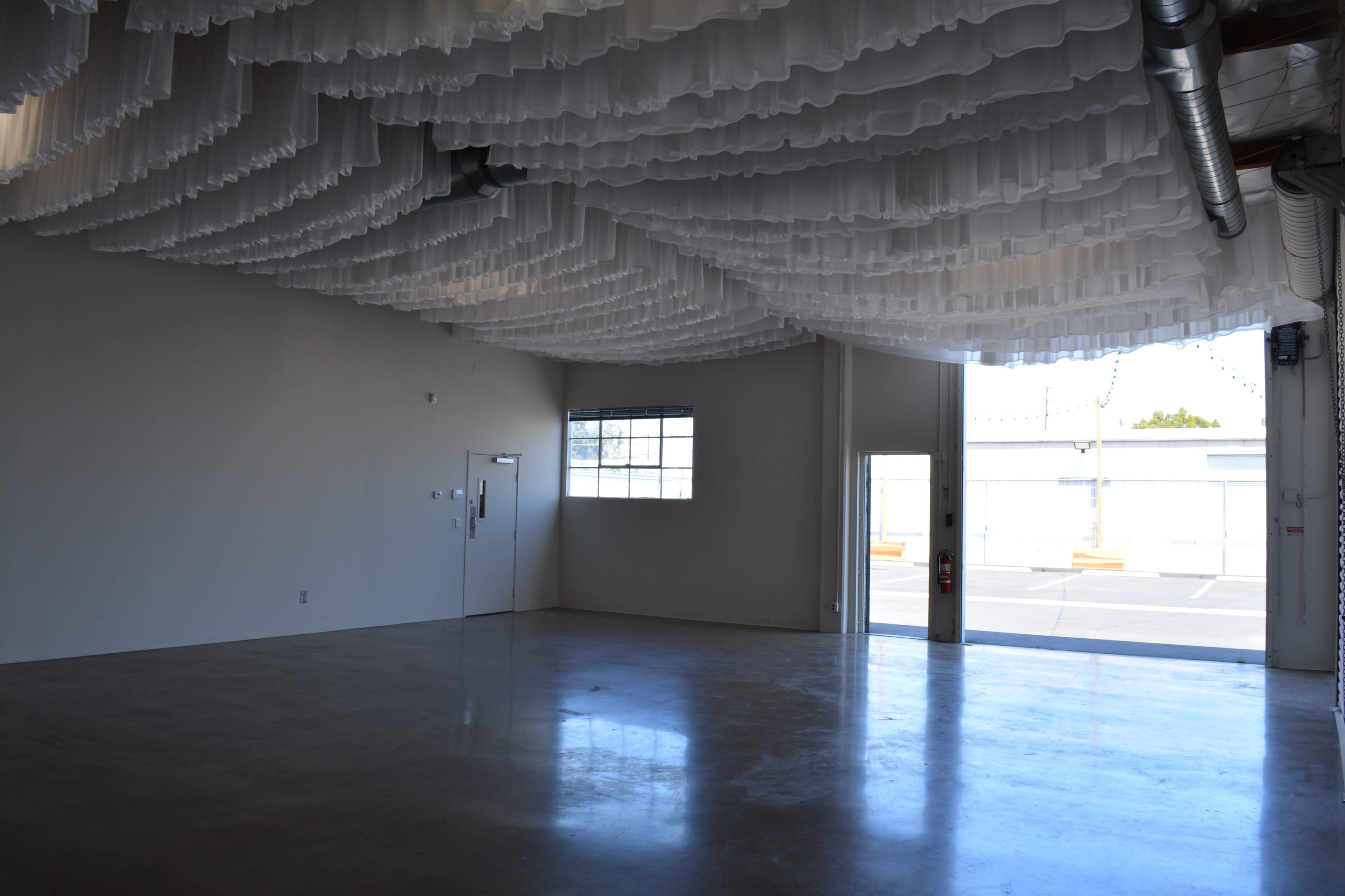 An empty industrial space with a ceiling decorated by layered white fabric and large windows allowing natural light.