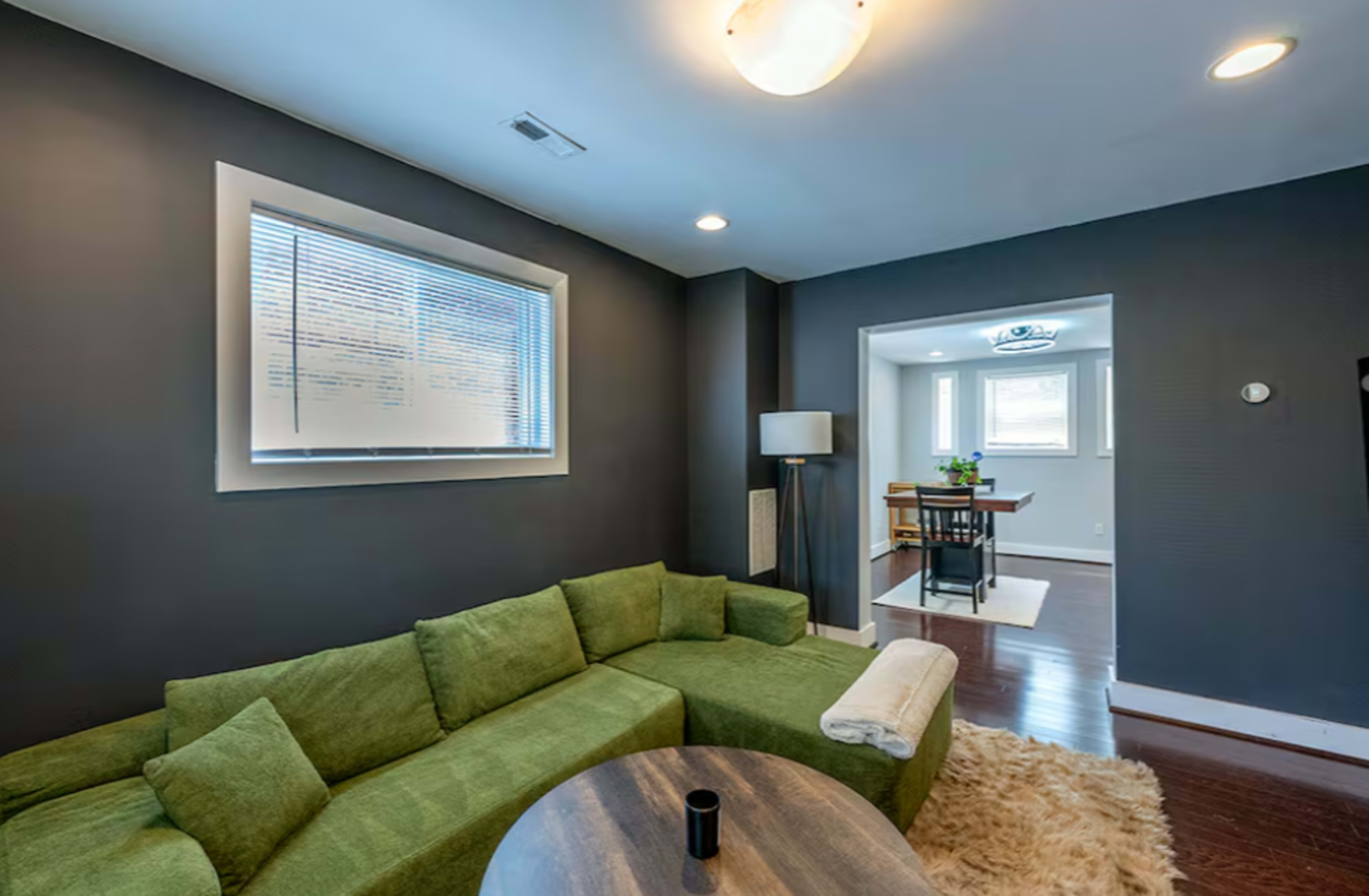 A green sectional sofa is positioned in a dimly lit living room with dark walls and a window, leading to a bright dining area.