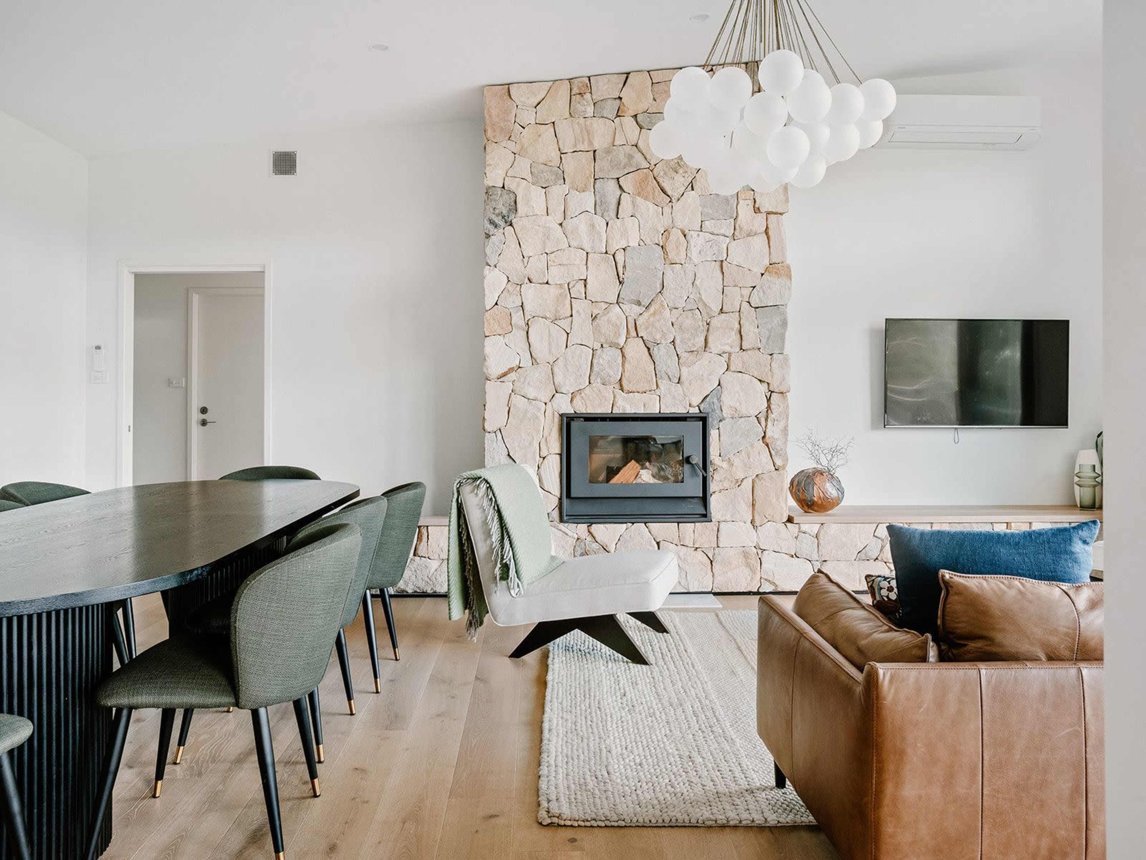 A modern living area with a stone fireplace, a large wooden dining table, green upholstered chairs, and a leather sofa.
