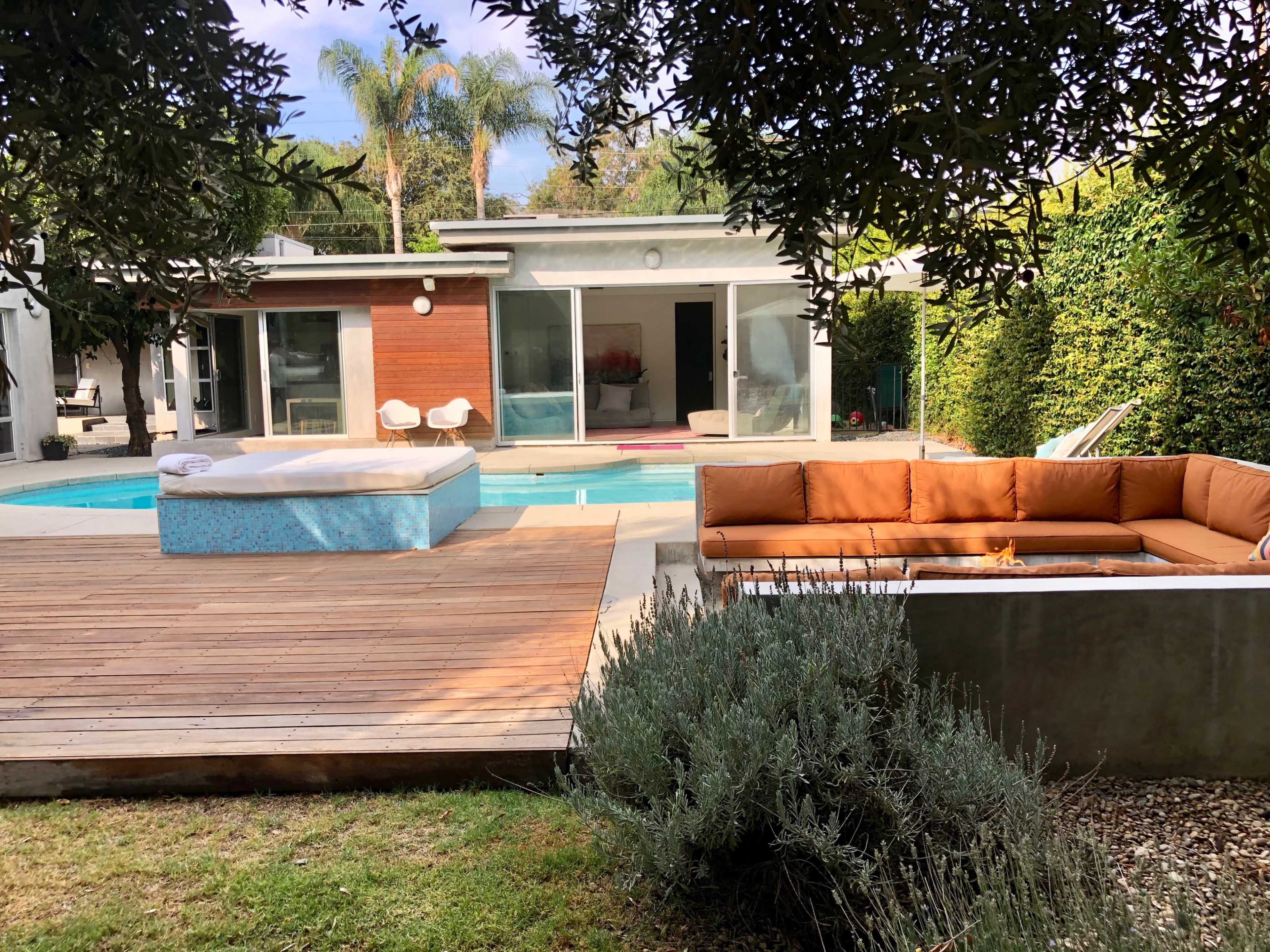 The image shows a modern backyard with a swimming pool, a hot tub, a wooden deck, and a spacious seating area surrounded by plants and trees.