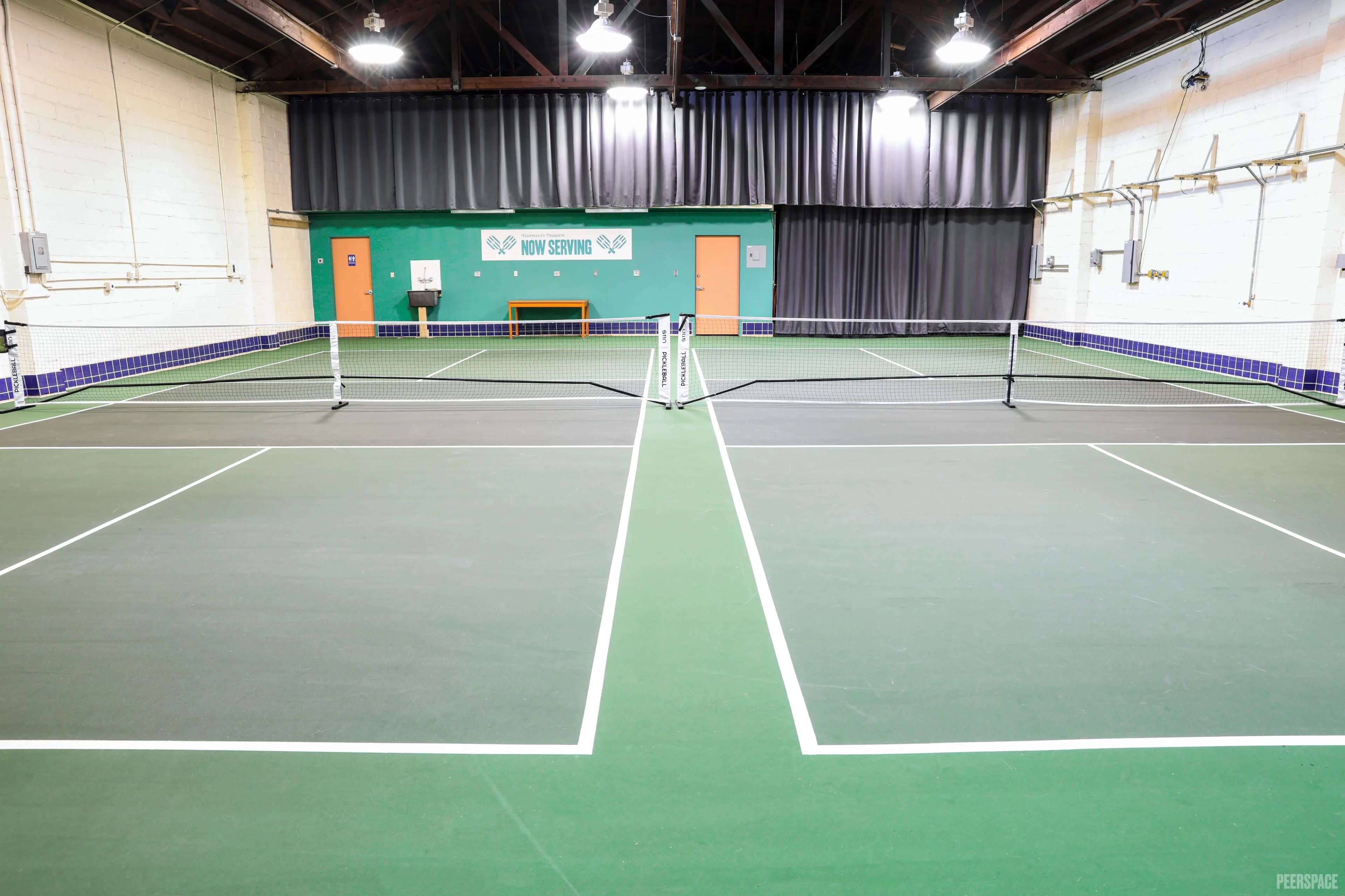 The image shows a sports facility with multiple indoor tennis courts, featuring green surfaces and nets in a well-lit environment.