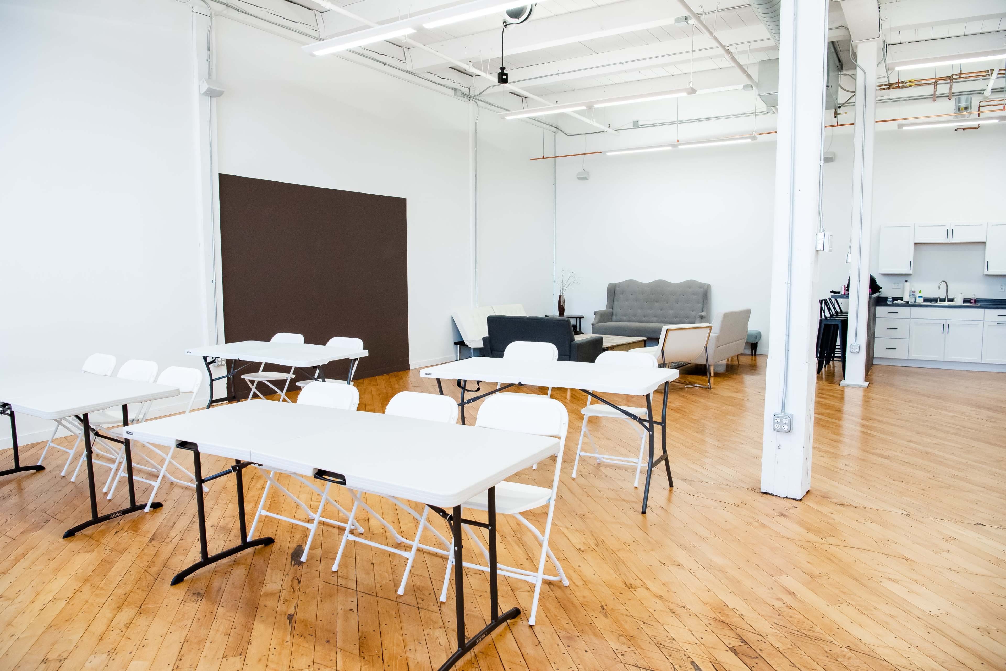 A spacious, well-lit room with several white folding tables and chairs arranged in a simple layout, alongside a seating area with a couch and a kitchenette in the background.