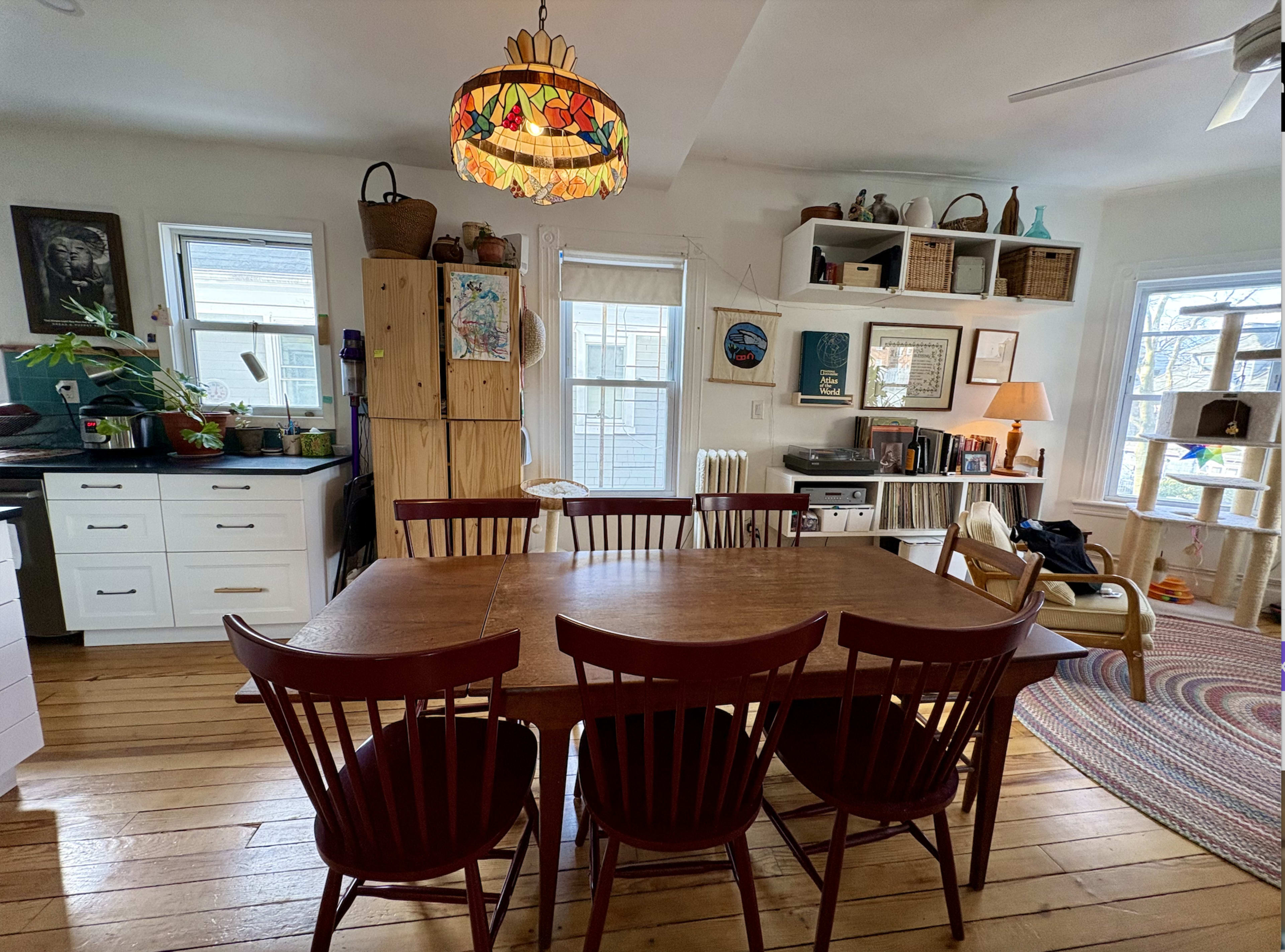 The image shows a cozy dining area with a wooden table surrounded by six chairs, a colorful hanging lamp, and a view of a kitchen and living space in the background.