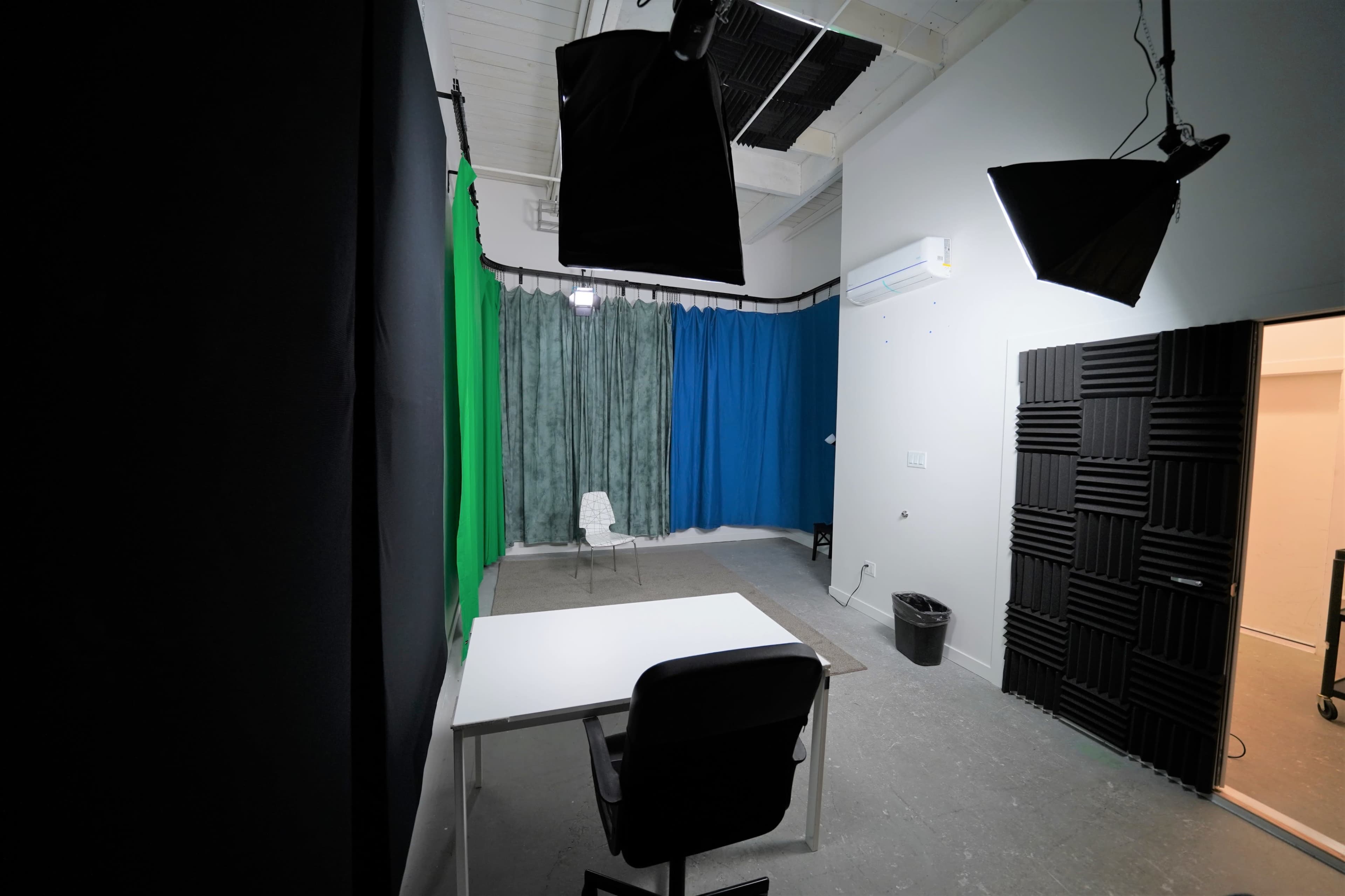 A blank studio room features a white table and chair, green and blue backdrops, and lighting equipment hanging from the ceiling.