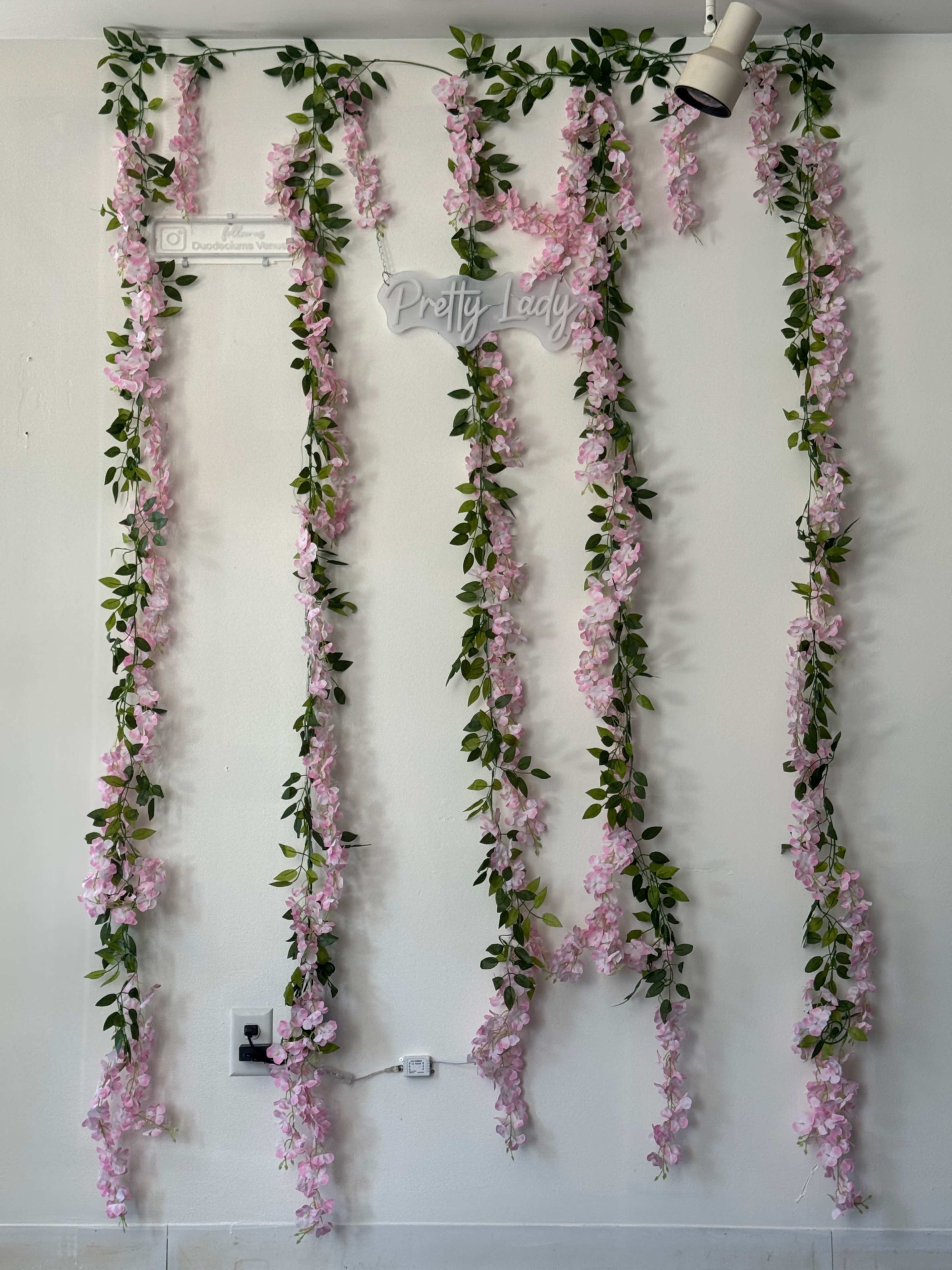 The image shows a wall decorated with vertical strands of pink flowers and green leaves, along with a sign that reads "Pretty Lady."