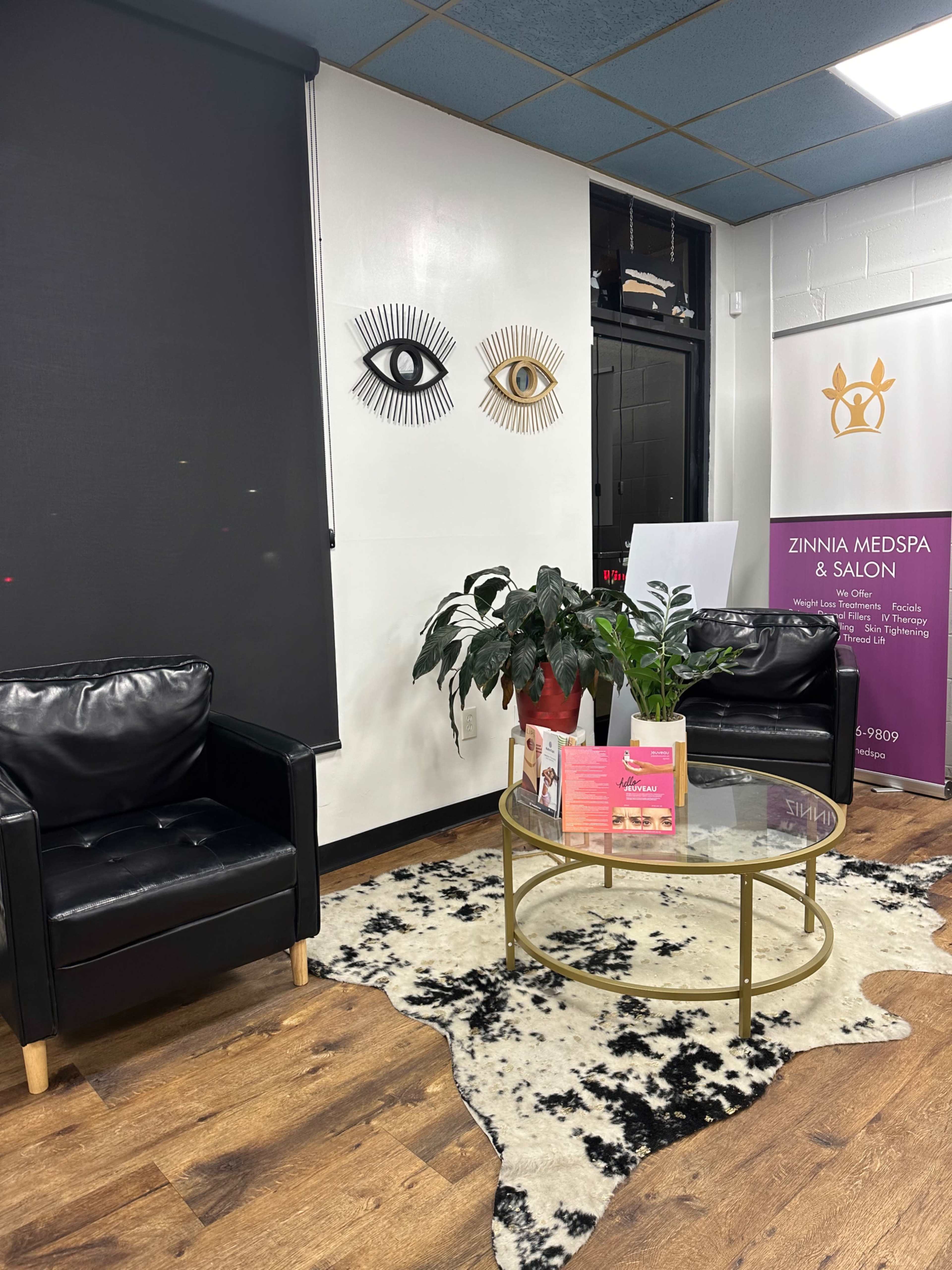 A modern salon interior with two black leather chairs, a round glass coffee table on a patterned rug, and a promotional display for "Zinnia Medspa & Salon."