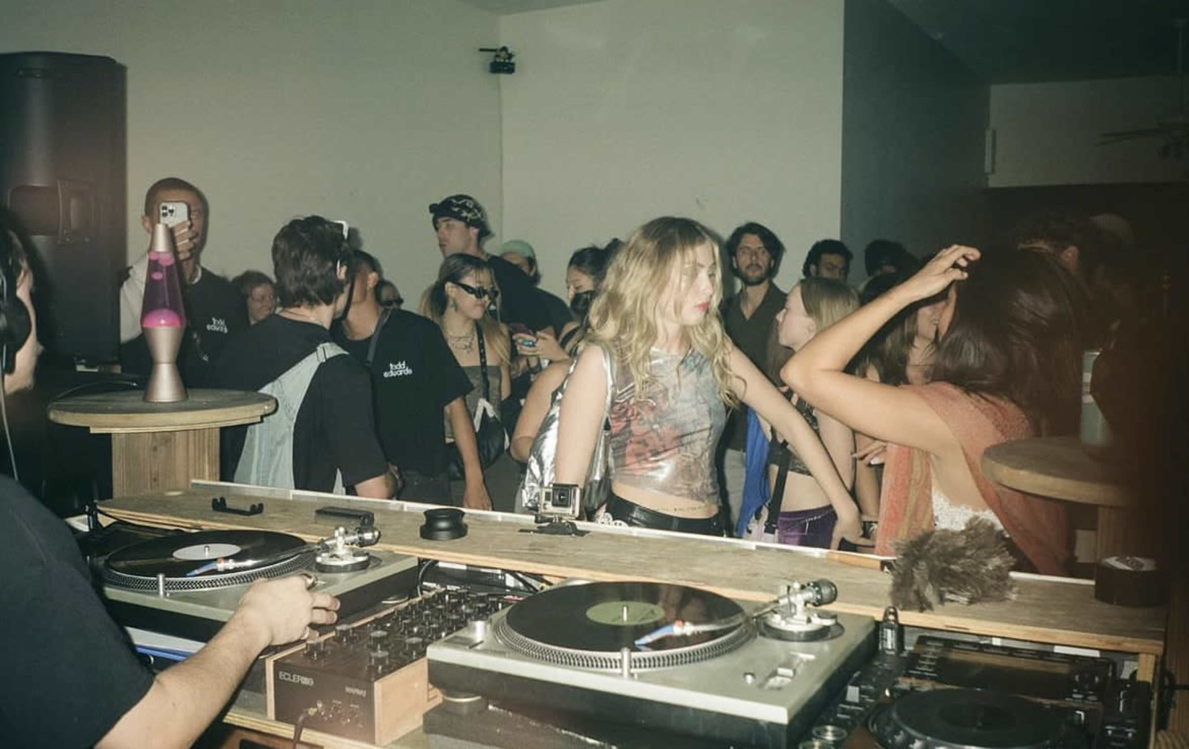 A crowd of people dances in a dimly lit room while a DJ plays records from a table surrounded by turntables.