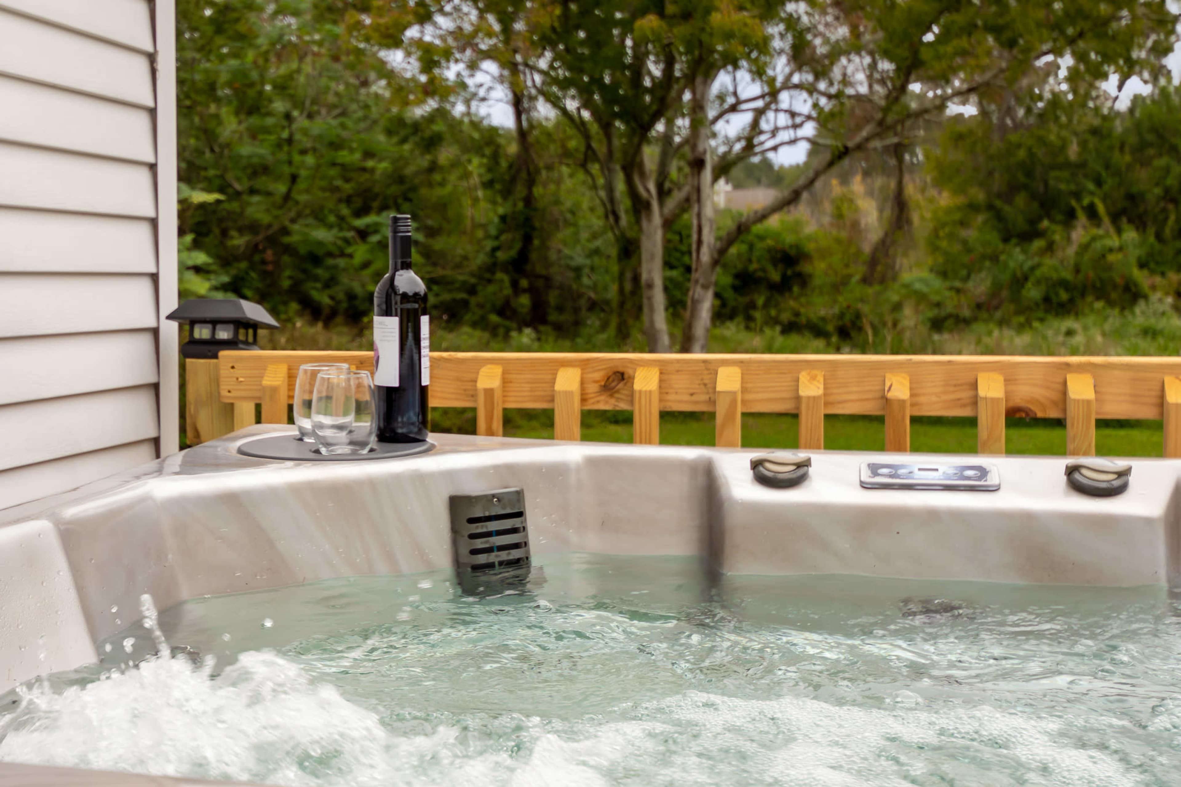 A hot tub with water splashing is positioned next to a wooden fence, featuring a bottle of red wine and a glass.