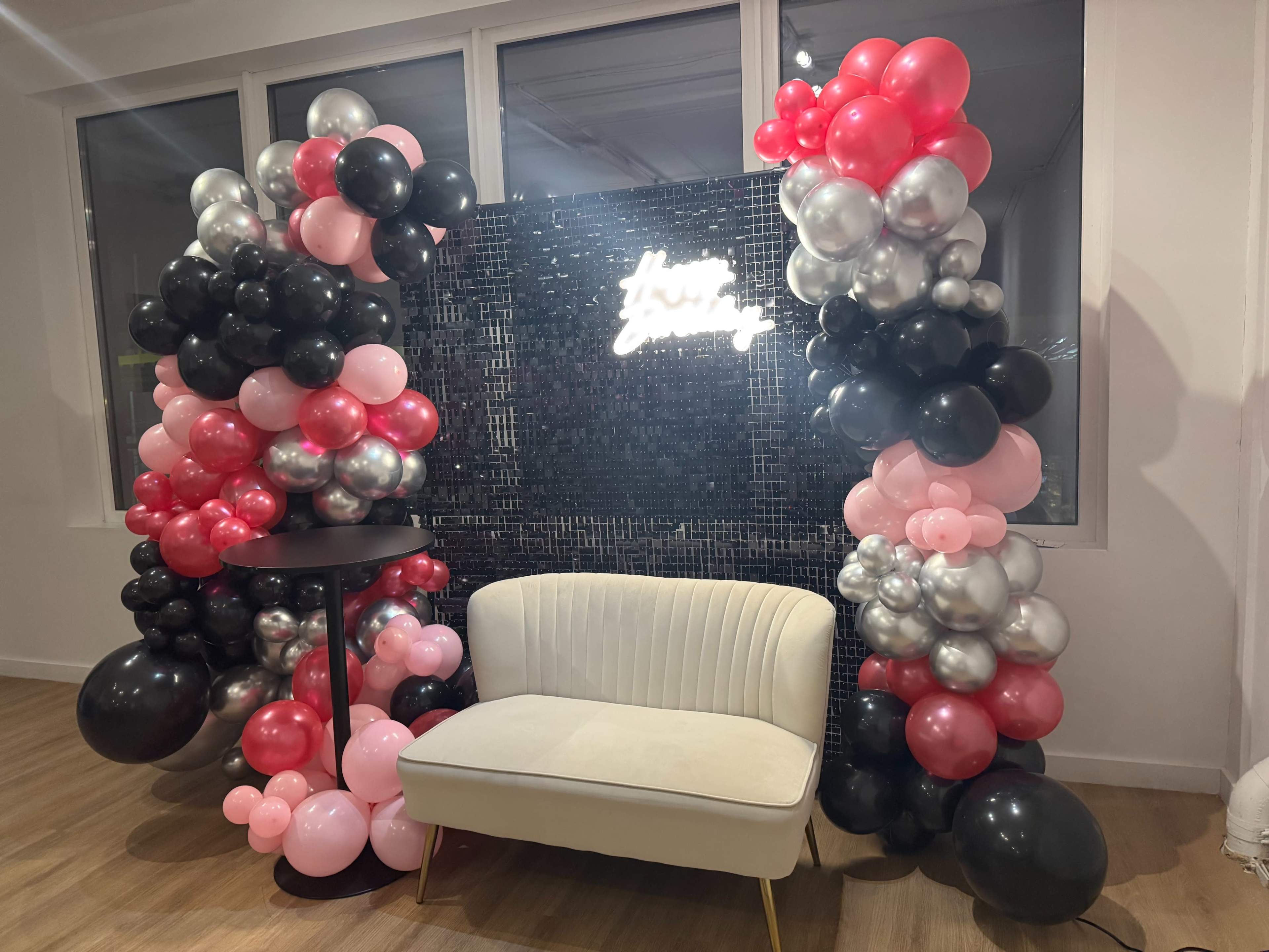 The image features a decorative backdrop made of balloons in black, pink, and silver, surrounding a stylish white couch and a small black table.