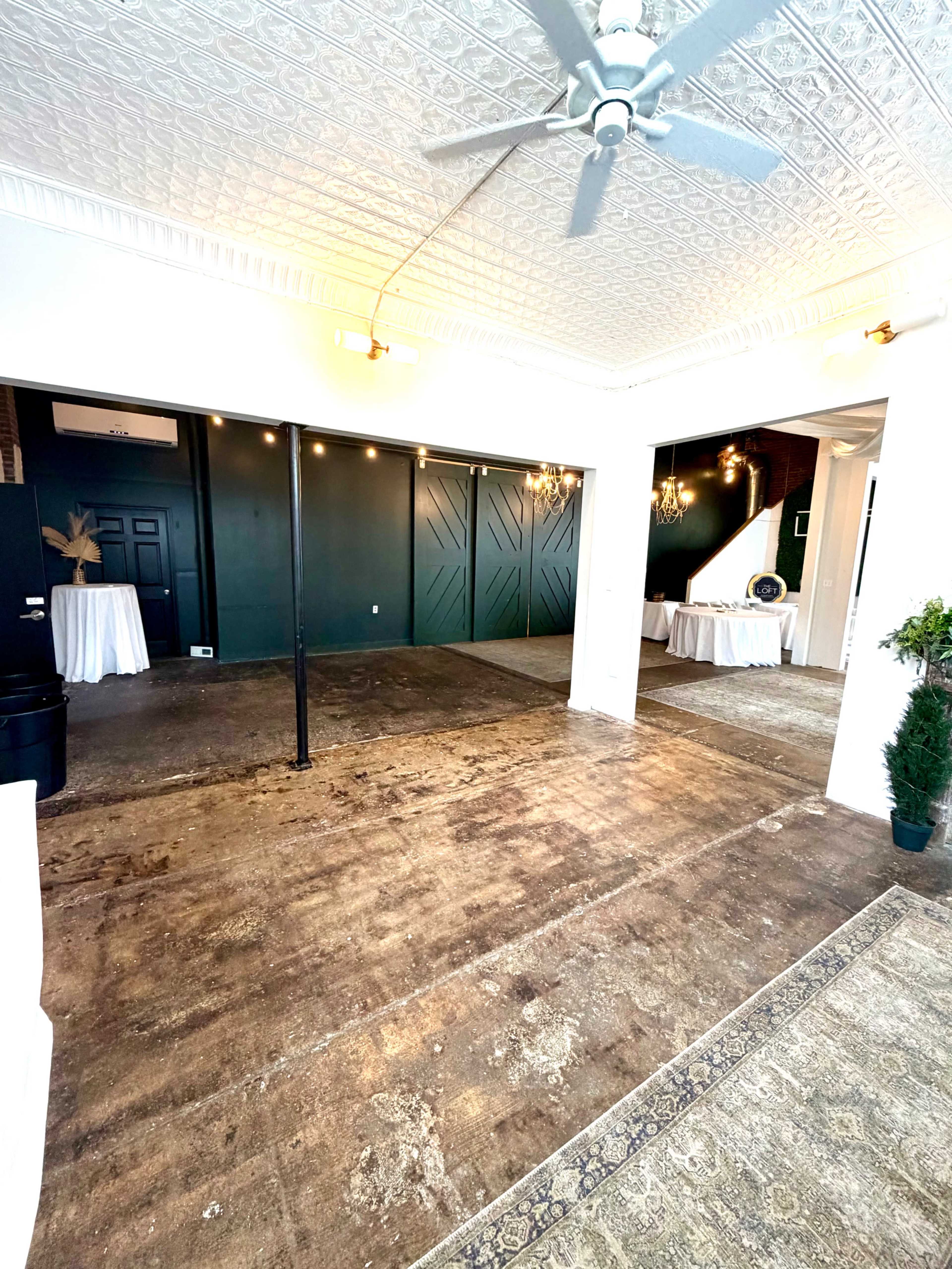The image shows a spacious room with a decorative ceiling, exposed wooden flooring, and a dark green accent wall, set up with tables draped in white cloths on one side.