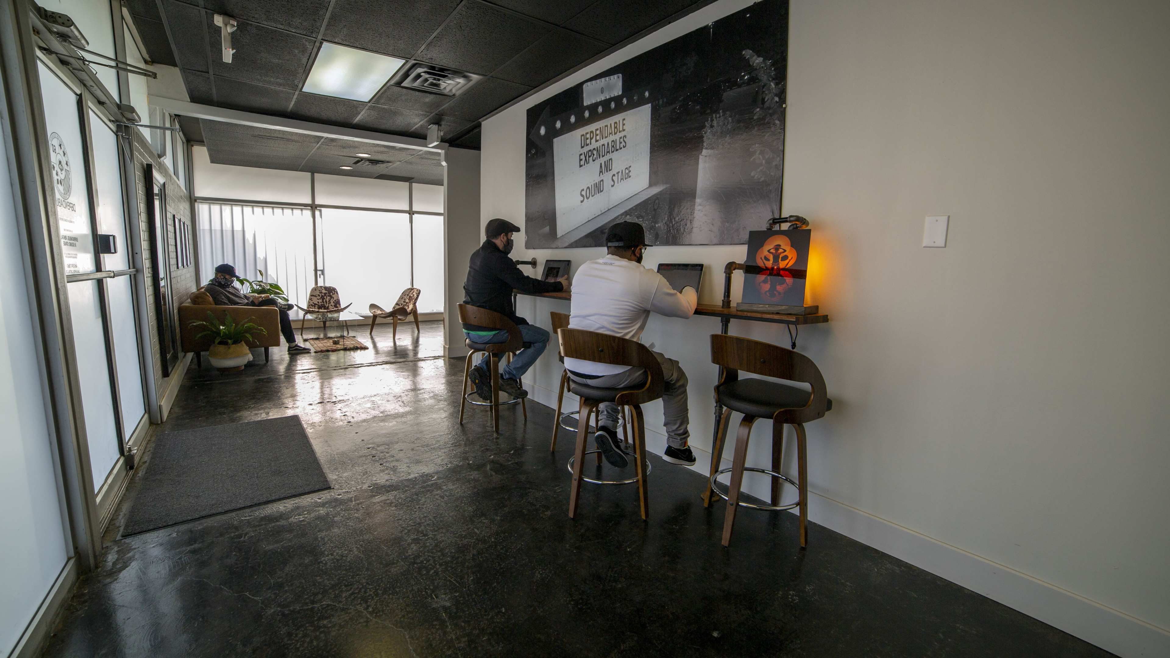 The image shows a modern interior space with two people sitting at high stools facing a wall with a large graphic, while another person sits in the background on a couch.