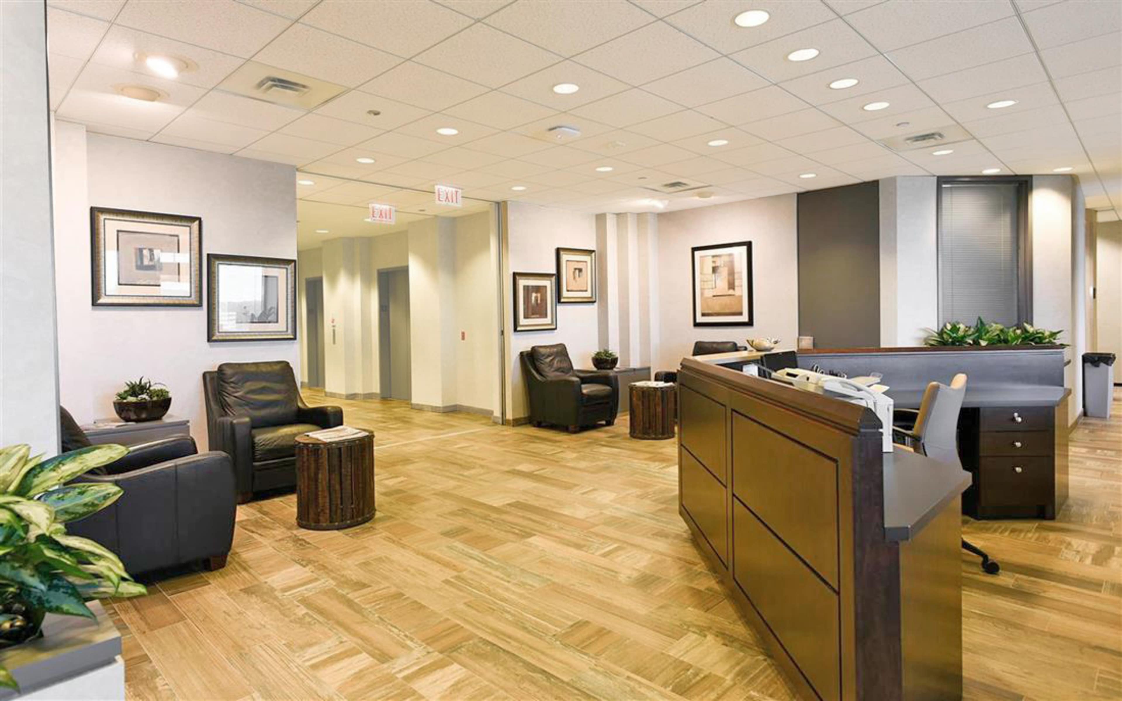 The image shows a modern waiting area with leather chairs, decorative plants, and a reception desk in an office environment.