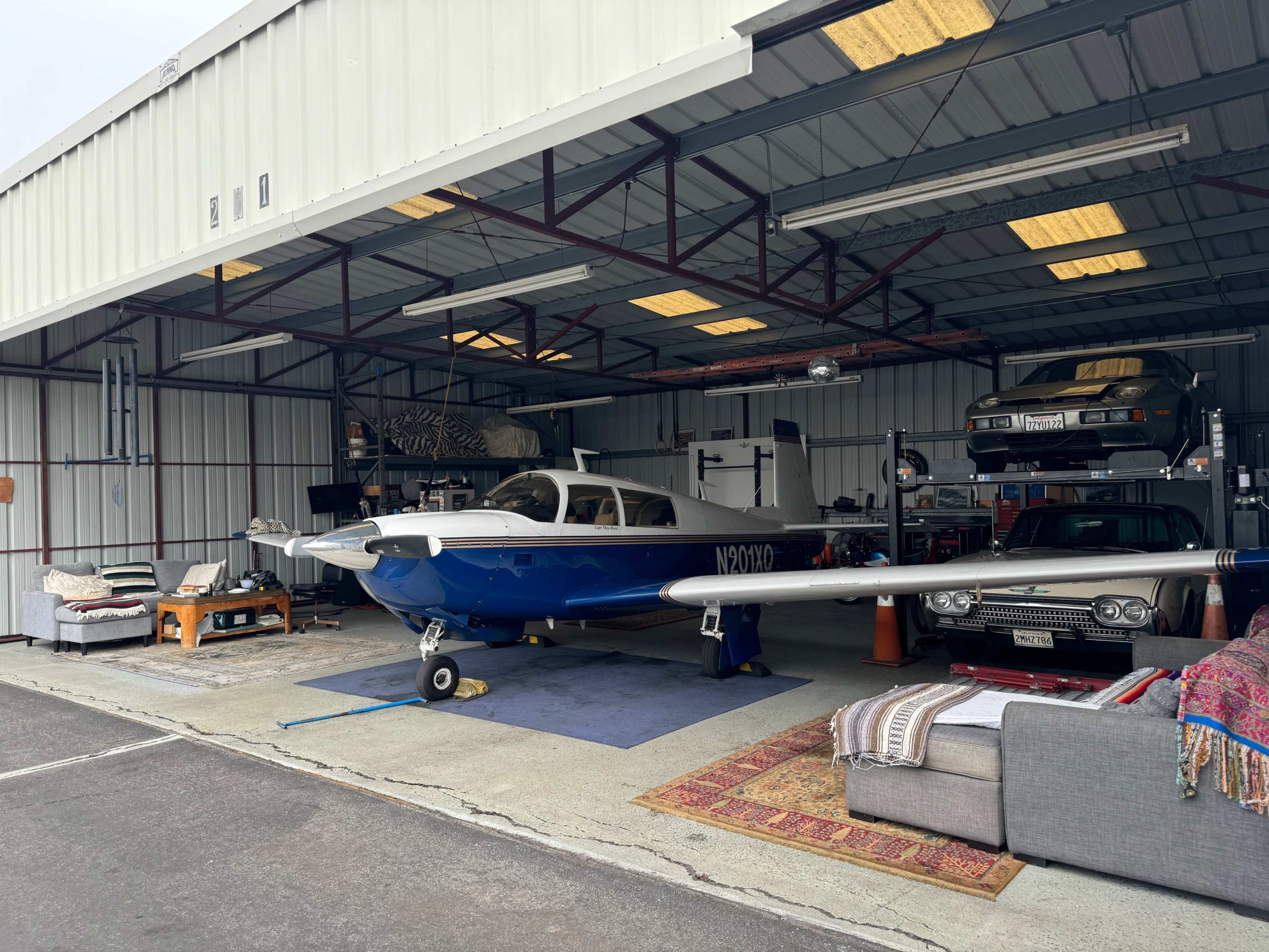 A blue and white airplane is parked inside a hangar alongside two vintage cars, with various furniture pieces arranged on the ground.