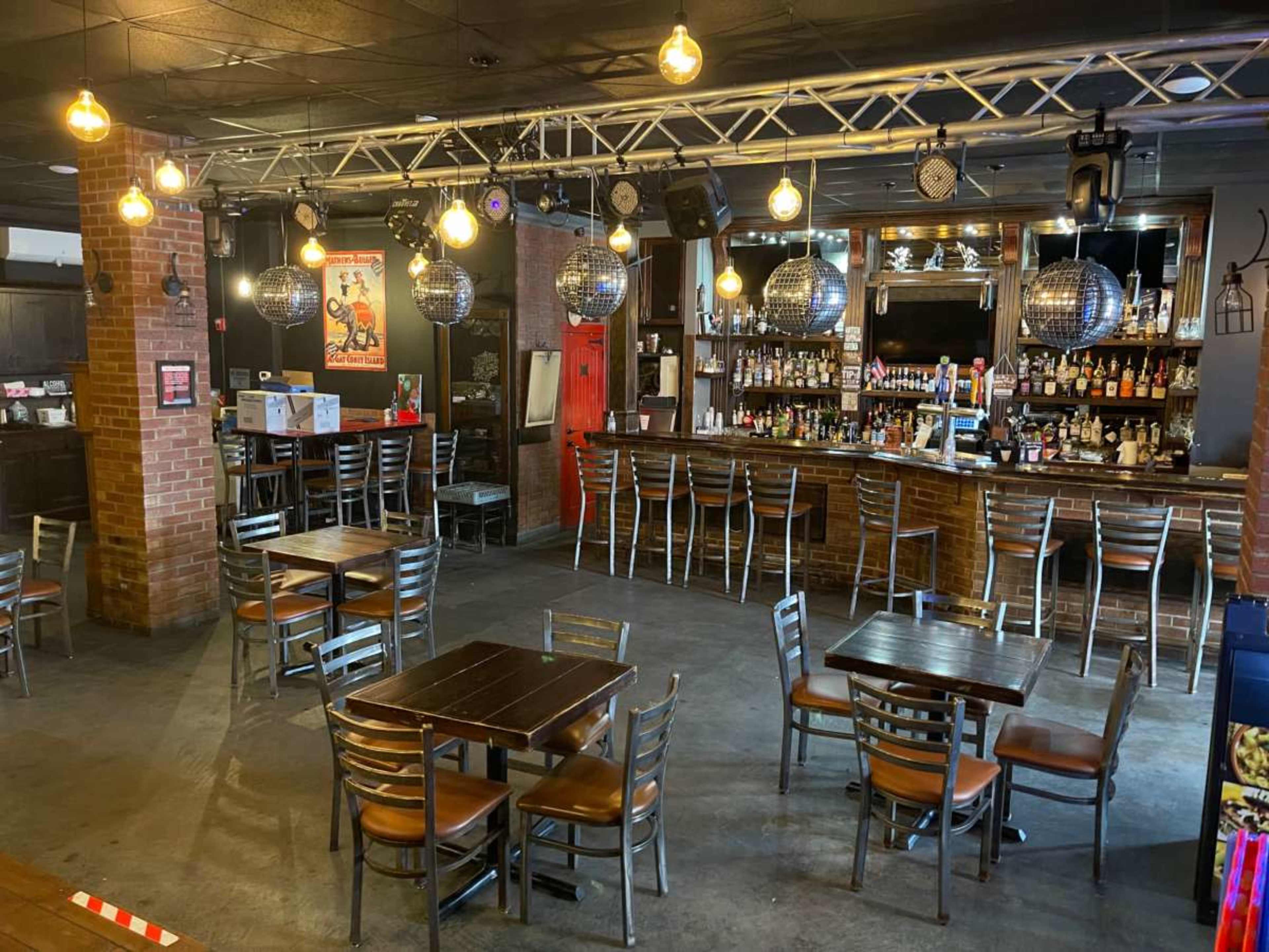 The image shows a spacious bar area featuring several tables and a well-stocked counter with various bottles, illuminated by pendant lights and disco balls overhead.