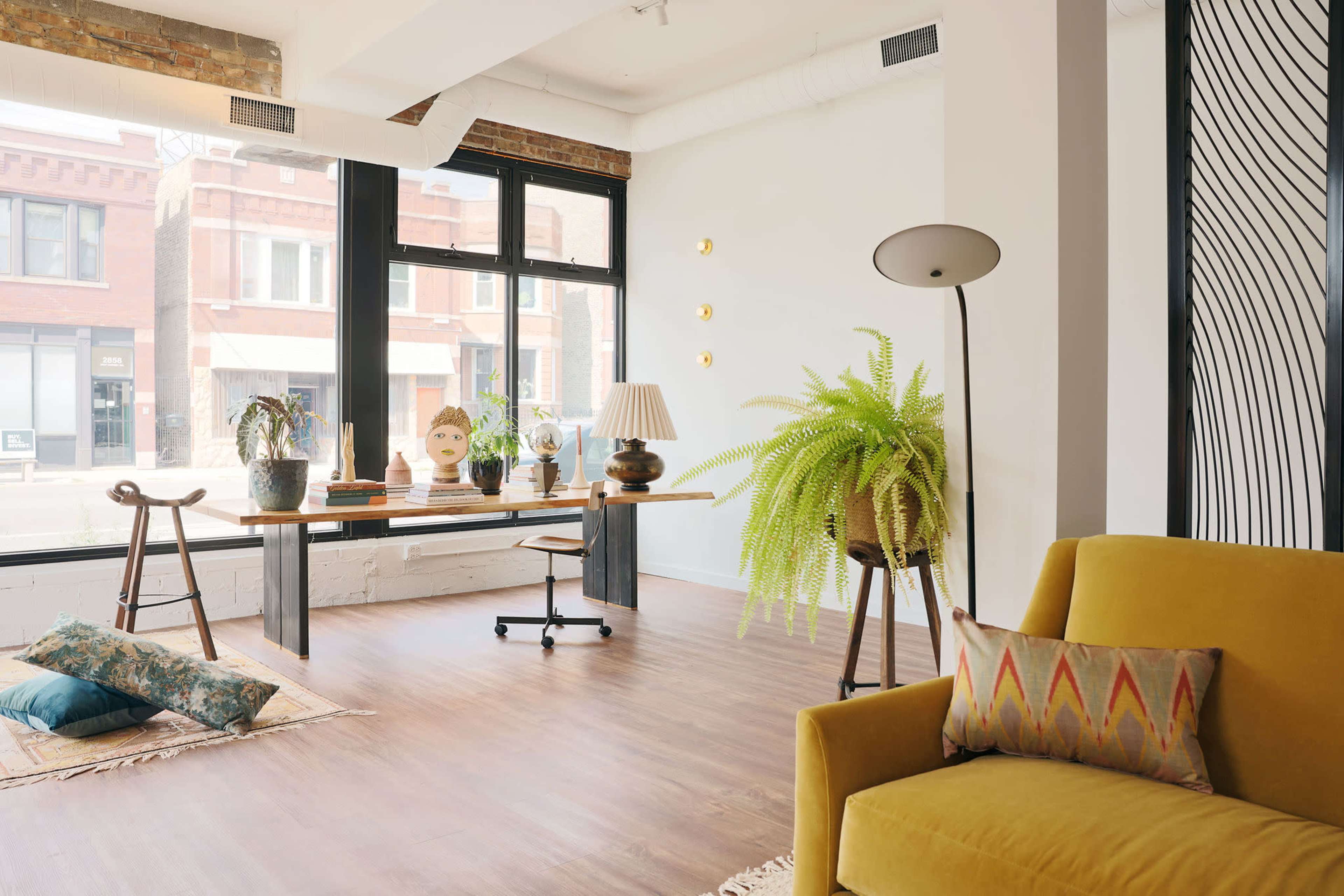 A modern office space features a desk near a large window, surrounded by plants, a floor lamp, and a mustard-colored sofa.