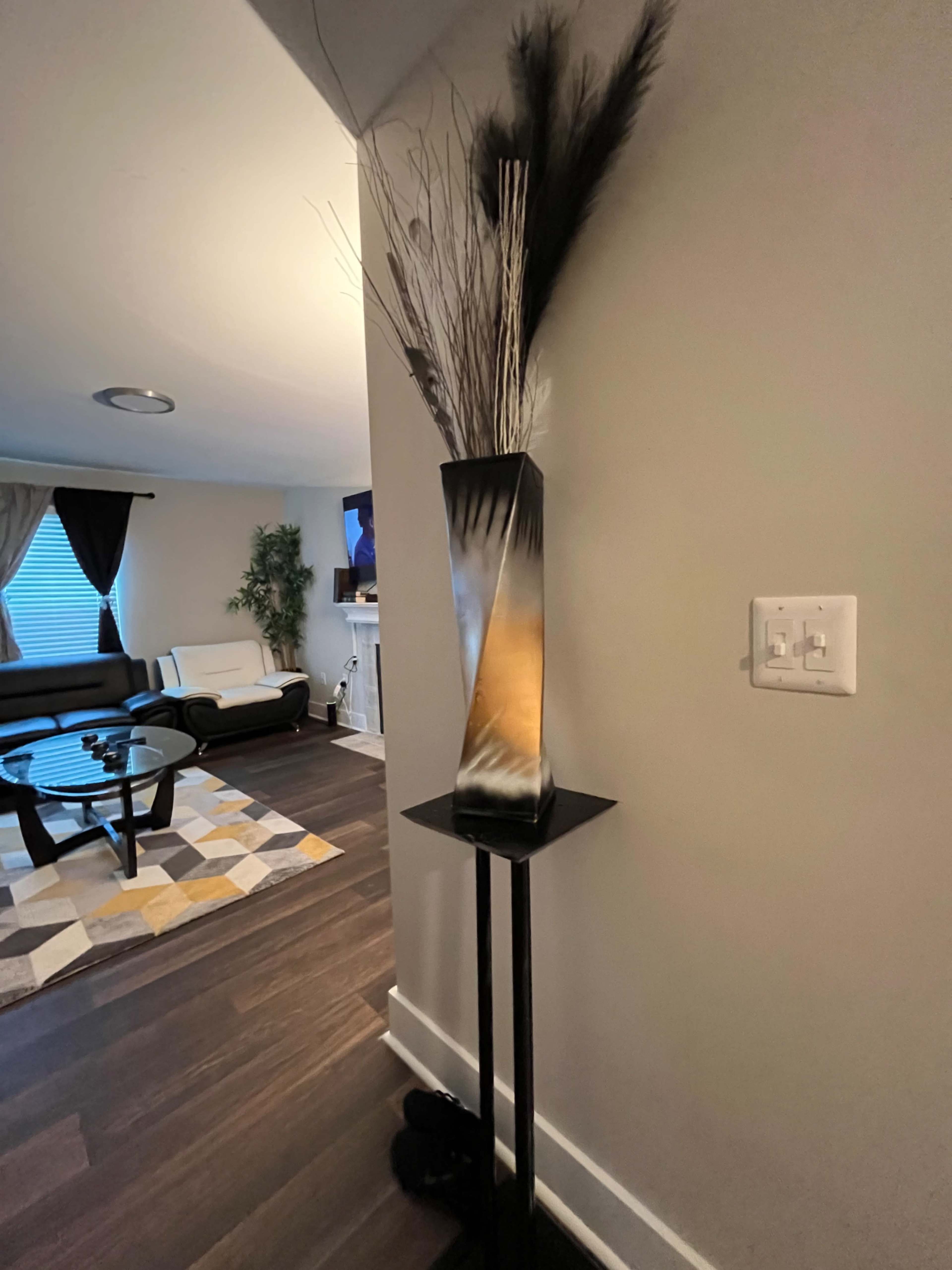 A tall, decorative floor vase with dried plants sits in a corner of a living room, next to a couch and a television.
