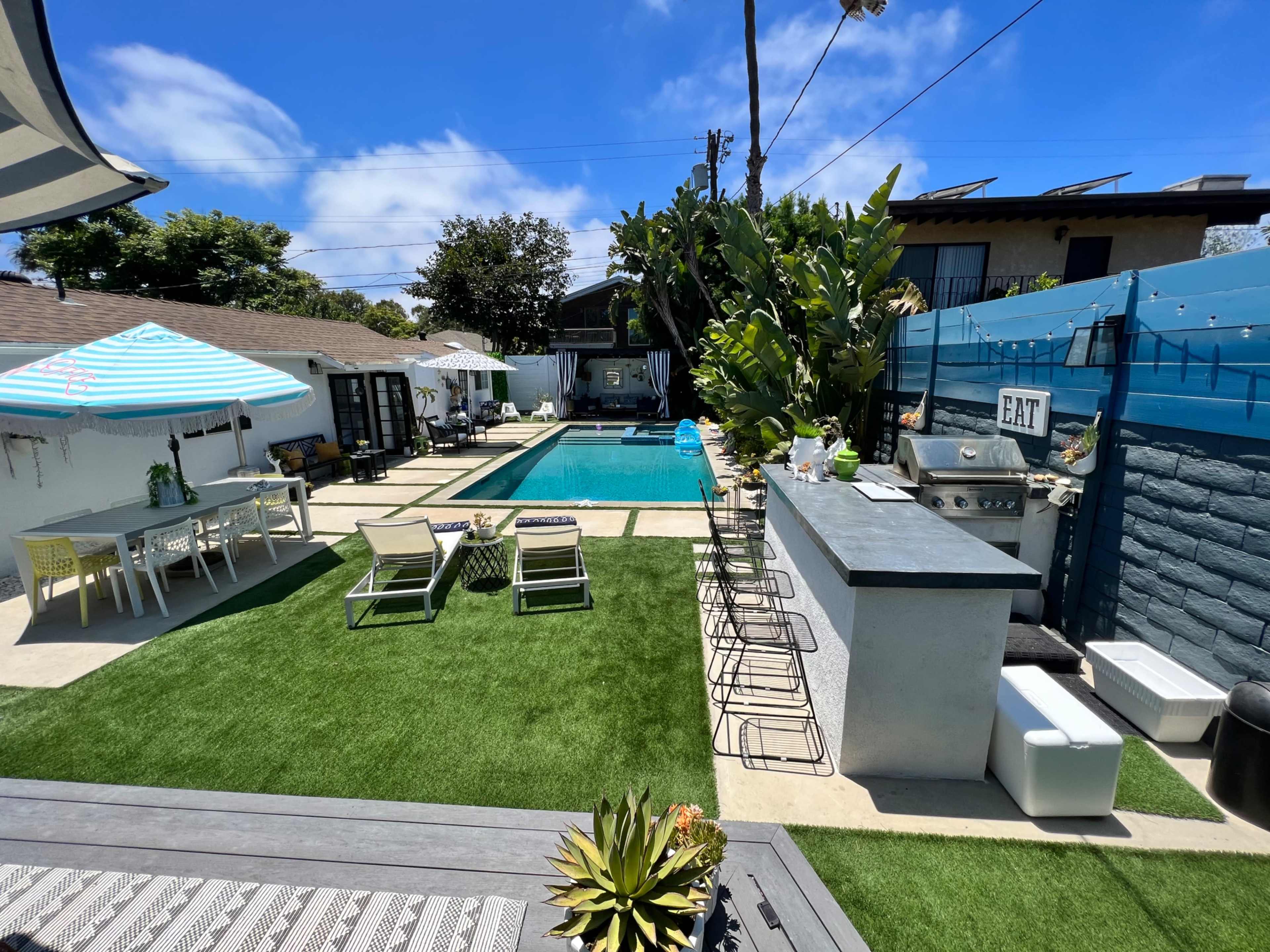 A modern outdoor space featuring a swimming pool, lounge chairs, a barbeque area, and several dining tables, all surrounded by greenery.