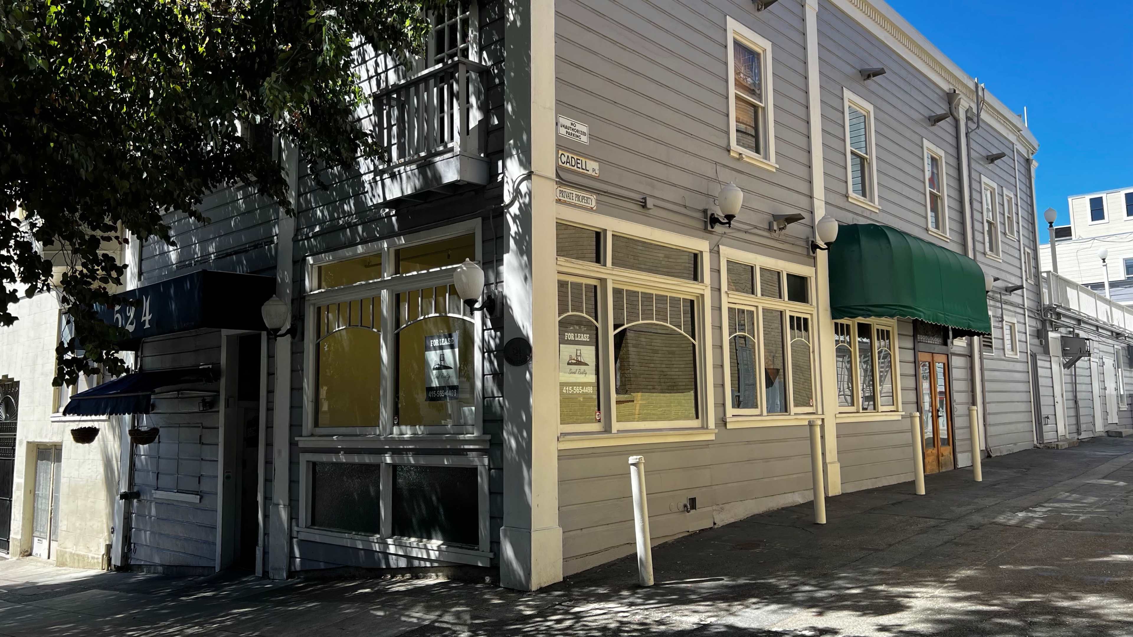 A gray building with large windows and an awning is situated on the corner of an urban street.