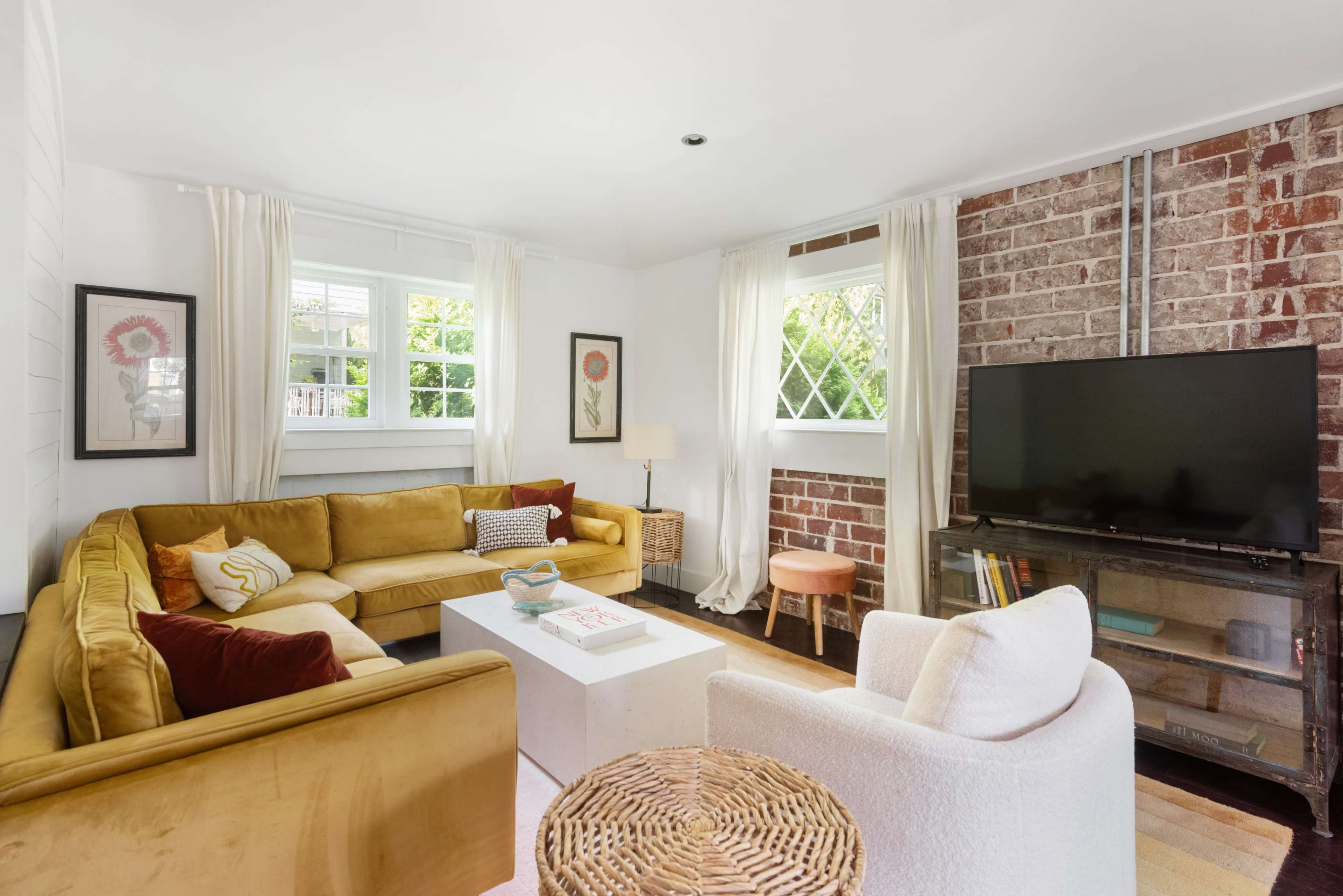 The living room features a yellow sectional sofa, a white coffee table, and a television on a stand against a brick wall, with large windows allowing natural light to enter.