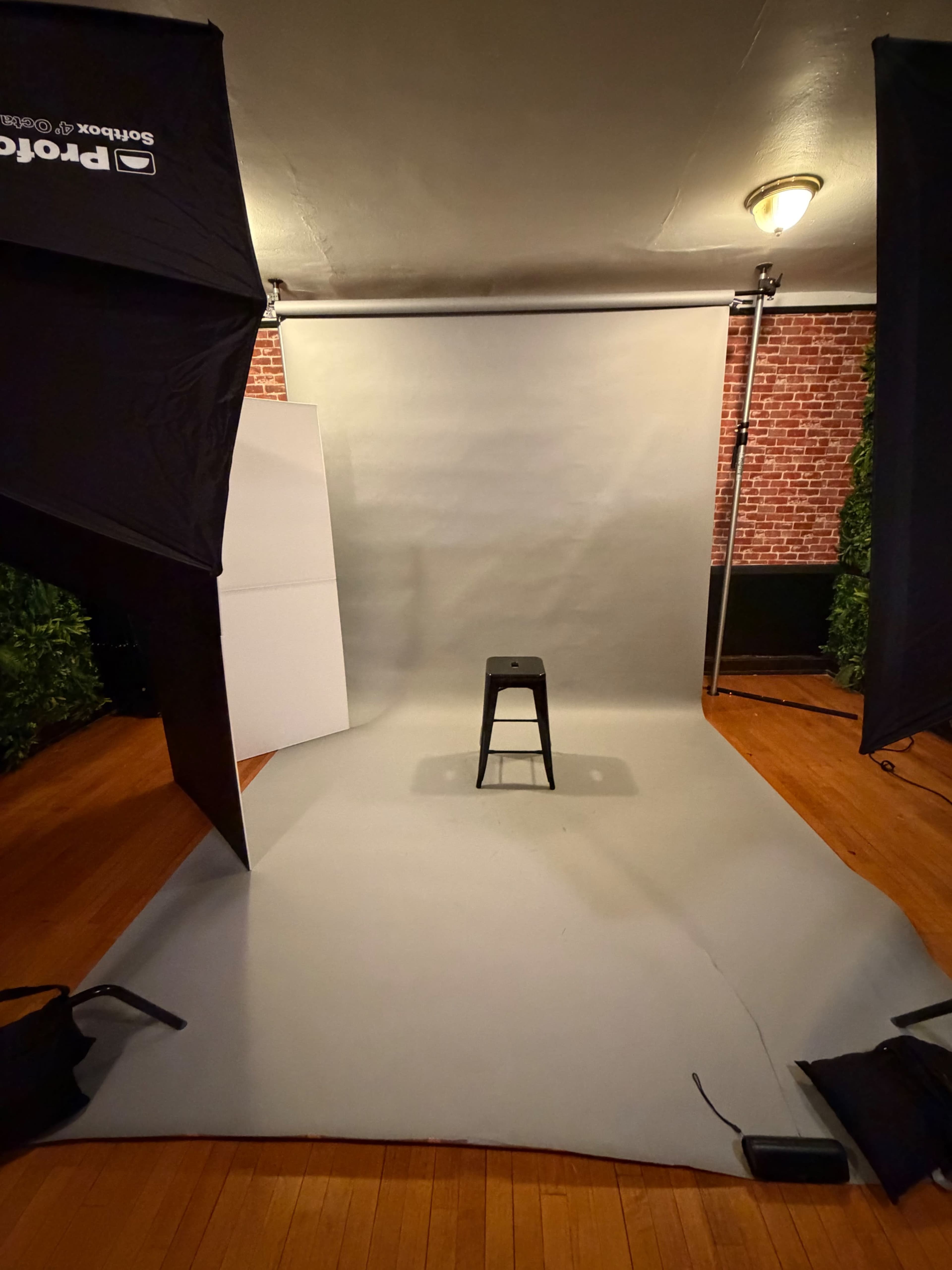 The image shows a photography studio setup with a gray backdrop, a black stool at the center, and lighting equipment on either side.