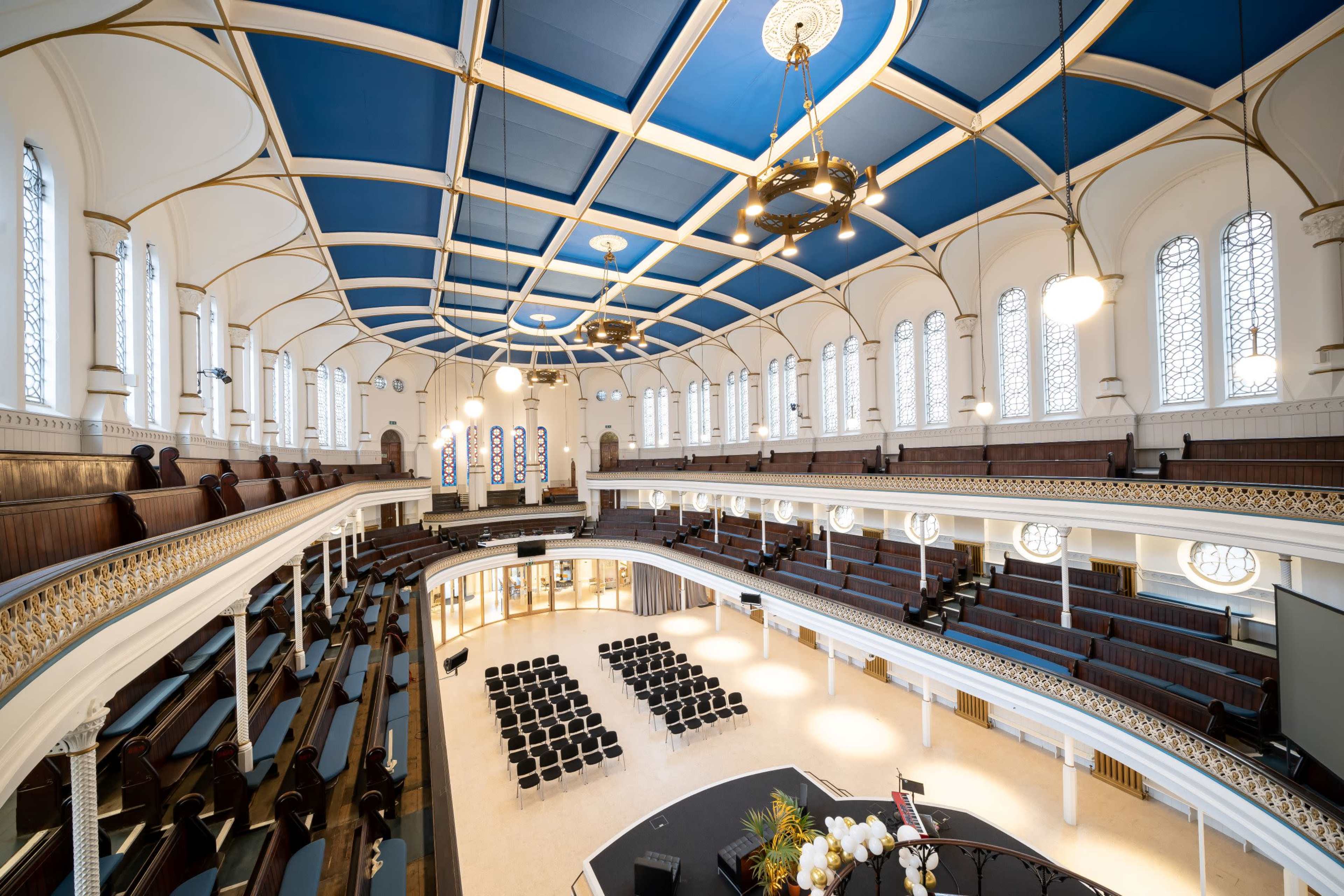 A spacious auditorium with a blue ceiling, multiple levels of seating, and large windows letting in natural light.