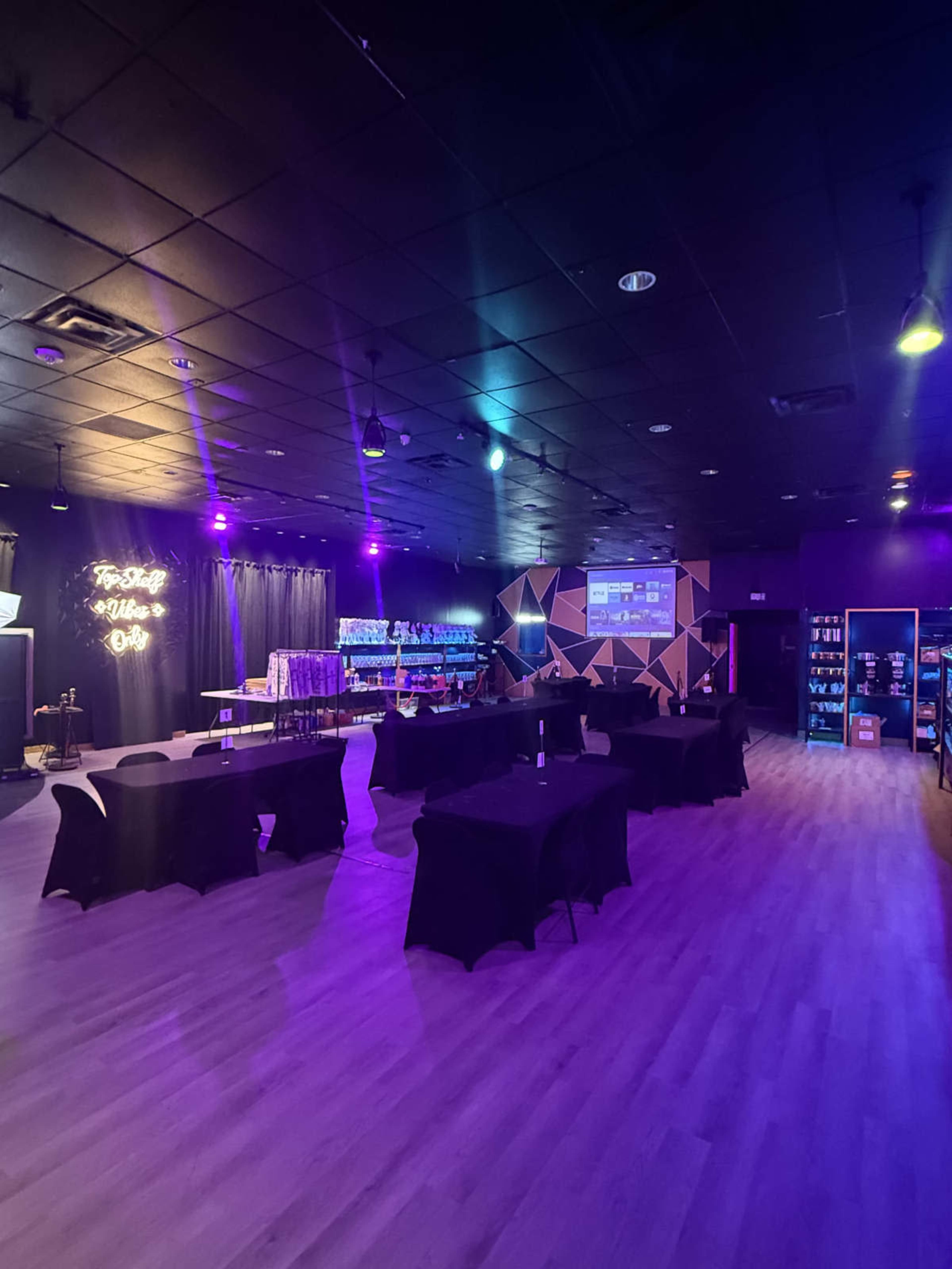 The image shows an event space with several round tables covered in black cloth, dimly lit with colorful lights, and decorated walls.