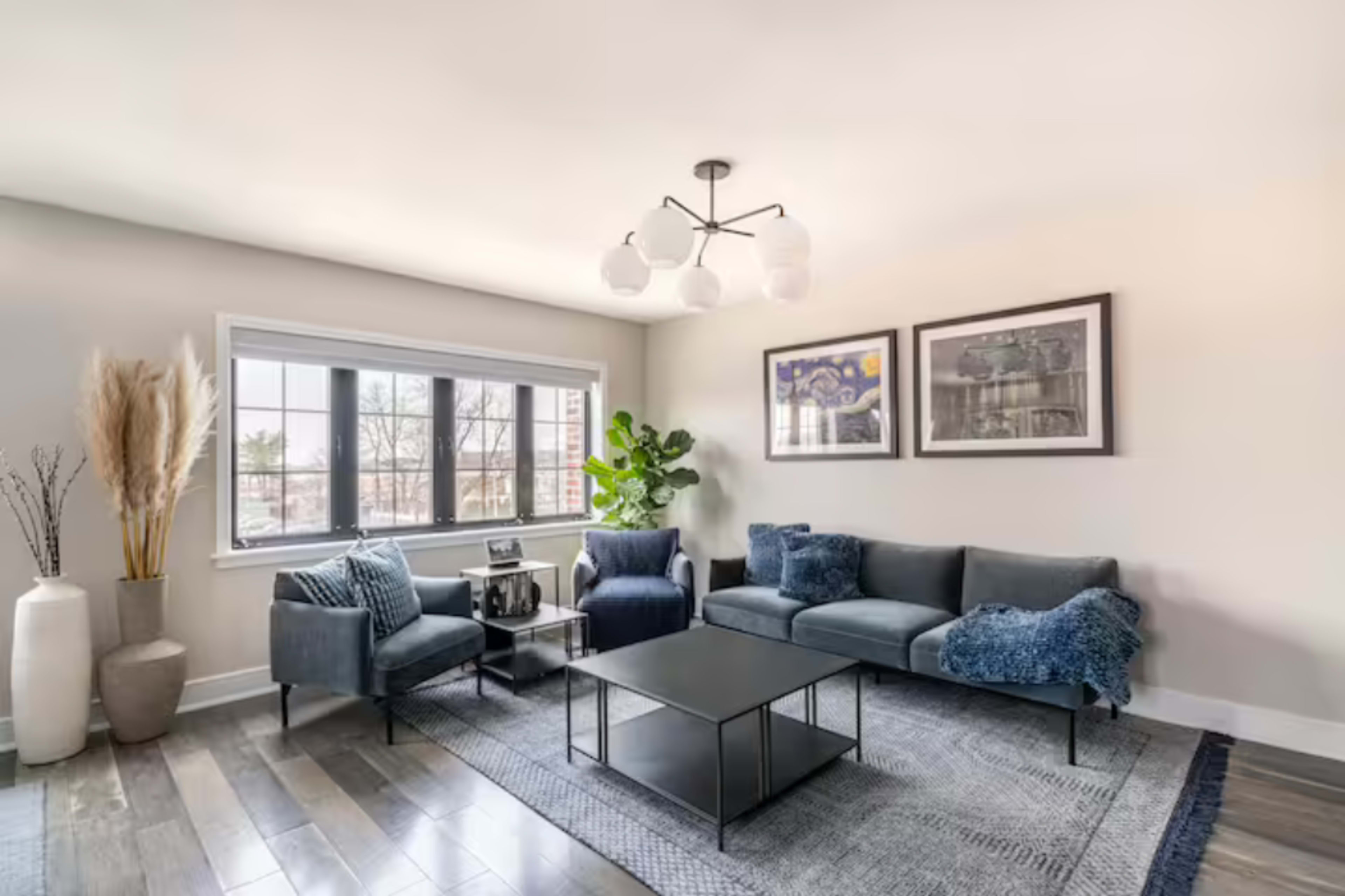 A modern living room with a dark sofa, a coffee table, framed artwork on the walls, and large windows allowing natural light to enter.