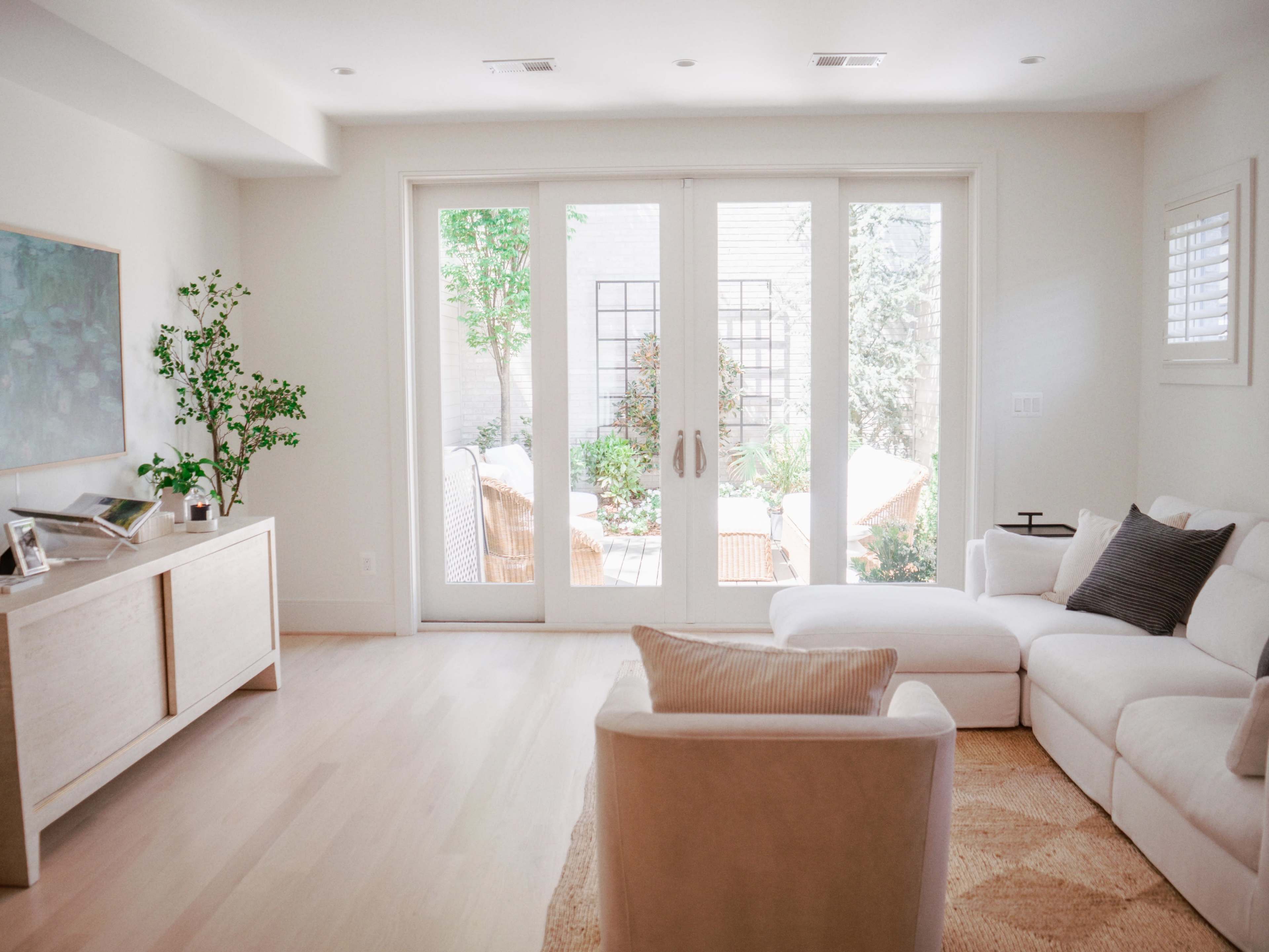 A bright, minimalist living room features a white sectional sofa, a light wood cabinet, and large glass doors opening to an outdoor space filled with greenery.