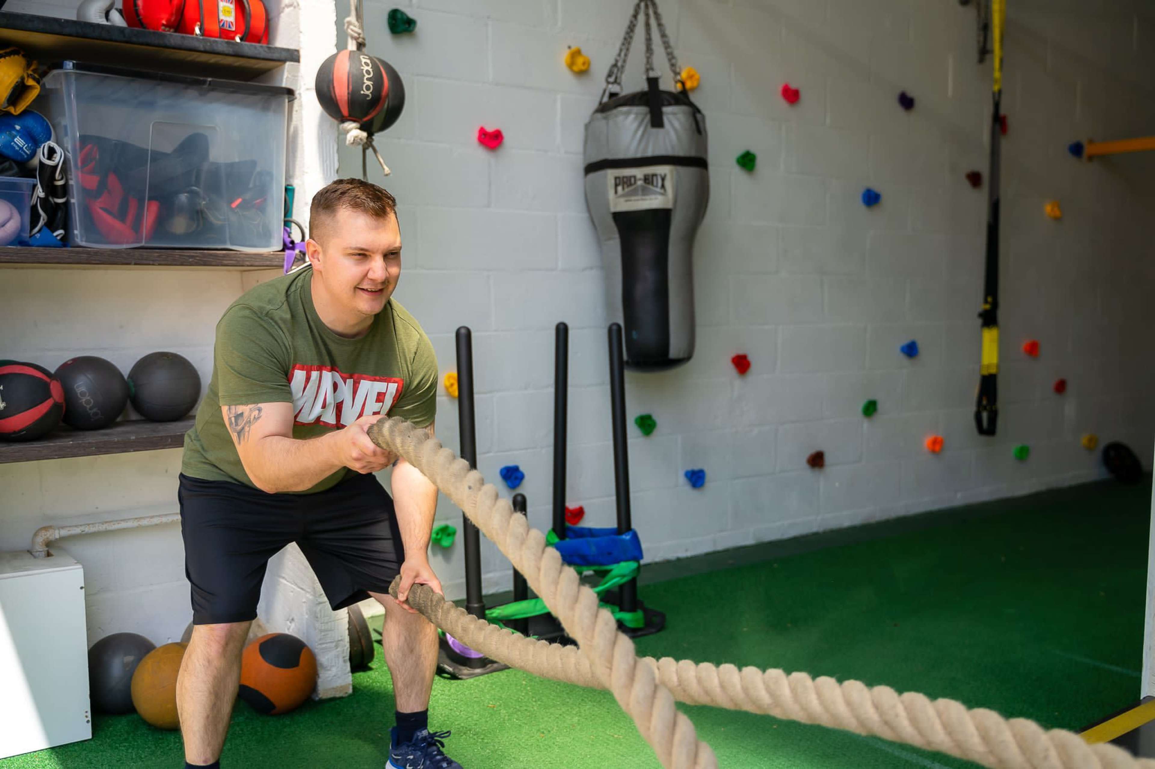 A man pulls battle ropes in a gym filled with exercise equipment and climbing holds on the wall.