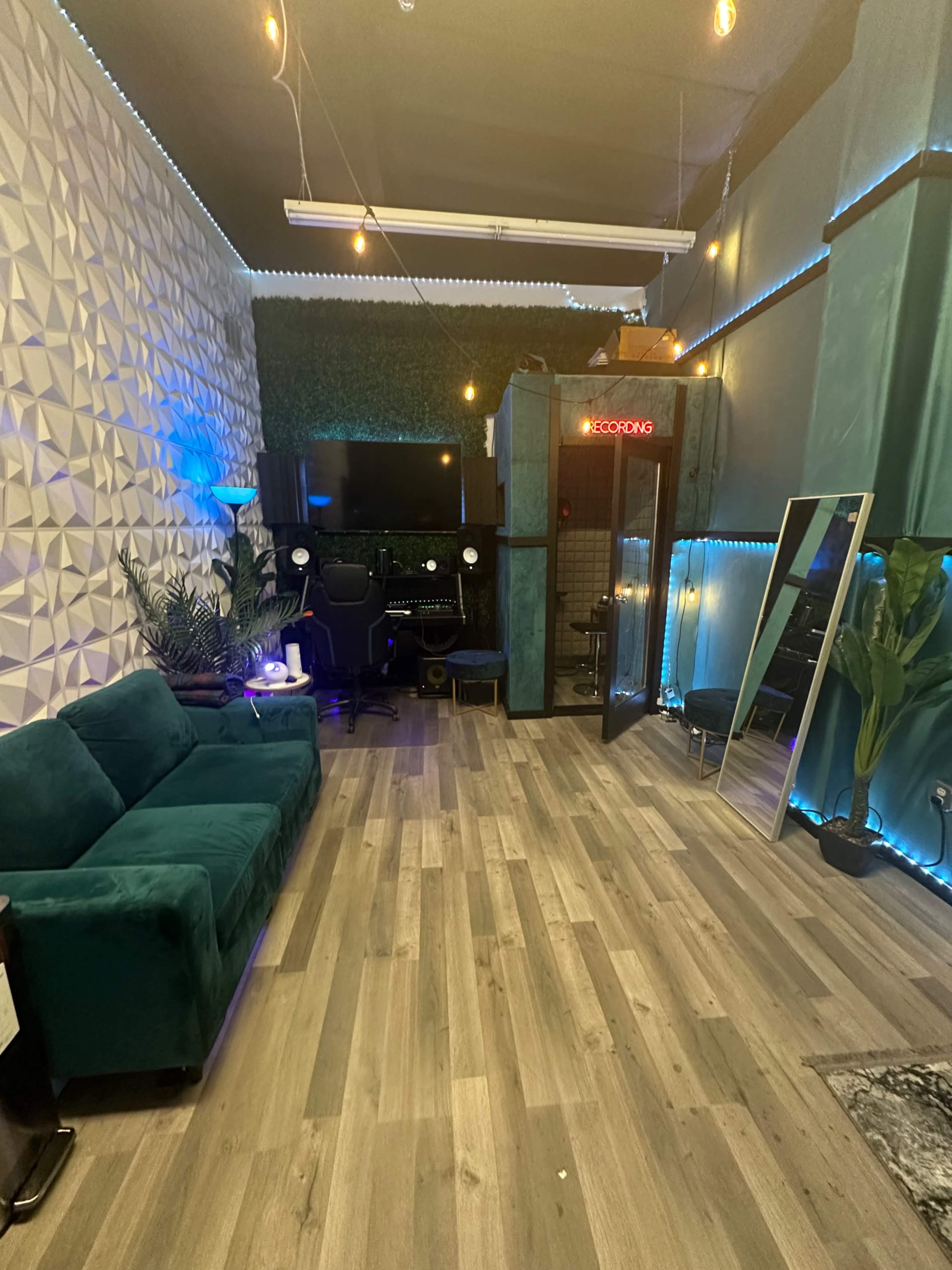 A modern recording studio featuring a teal couch, a workstation with a computer, and a soundproof booth enclosed with glass.