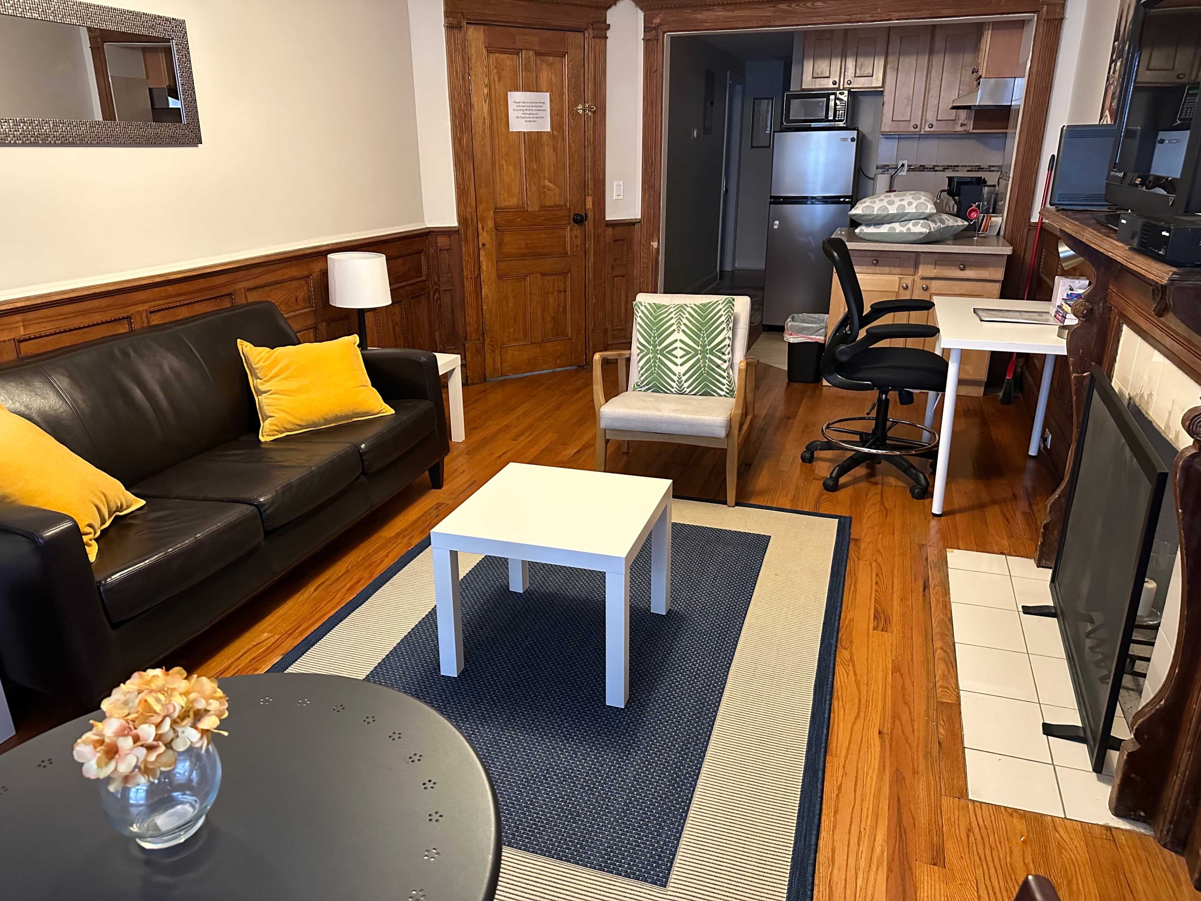 A cozy living room with a black leather sofa, a small white coffee table, and a dining area visible in the background.