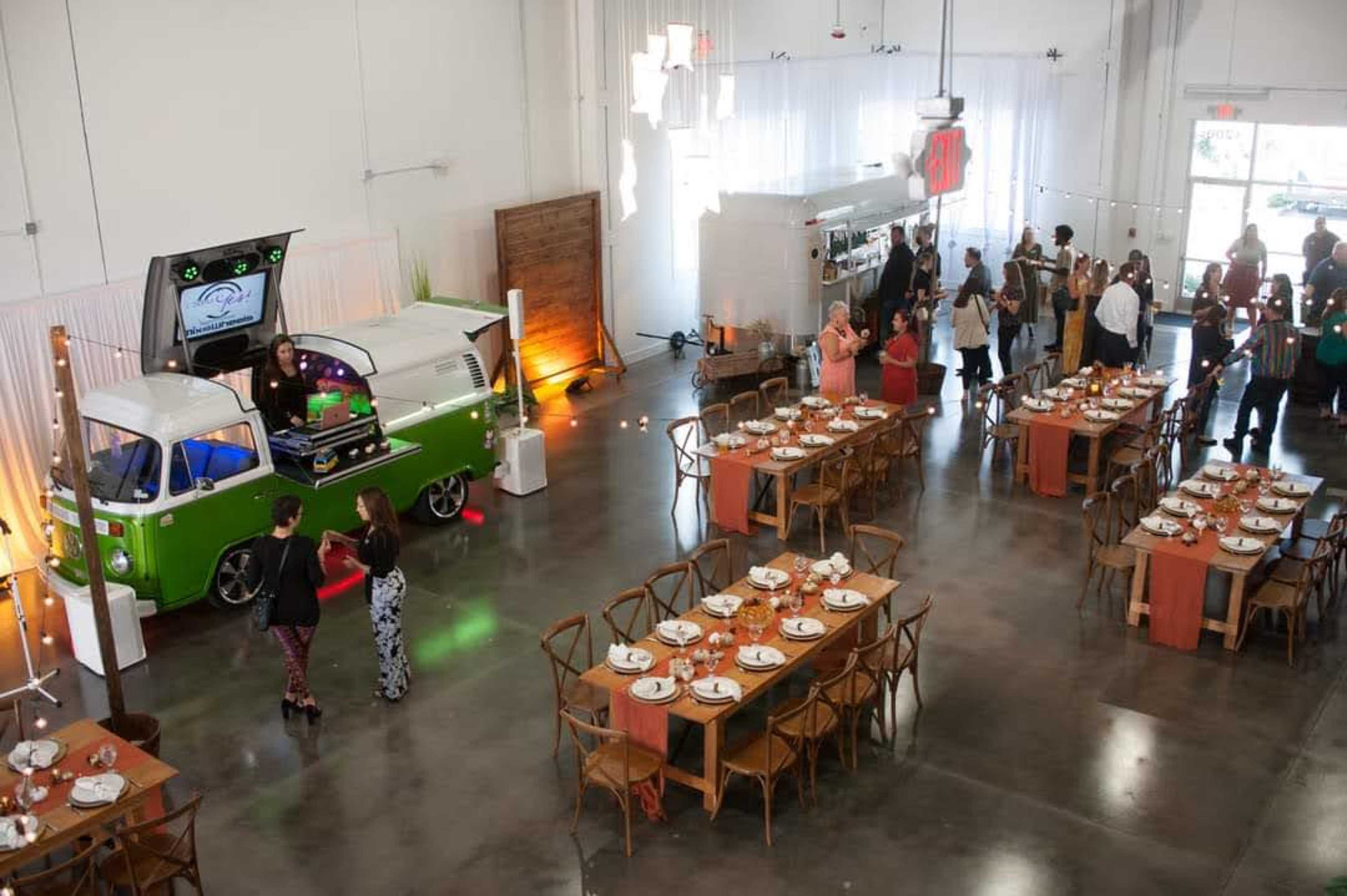 The image shows a spacious event venue with long wooden tables set for a meal, a DJ booth beside a vintage green van, and guests mingling in the background.