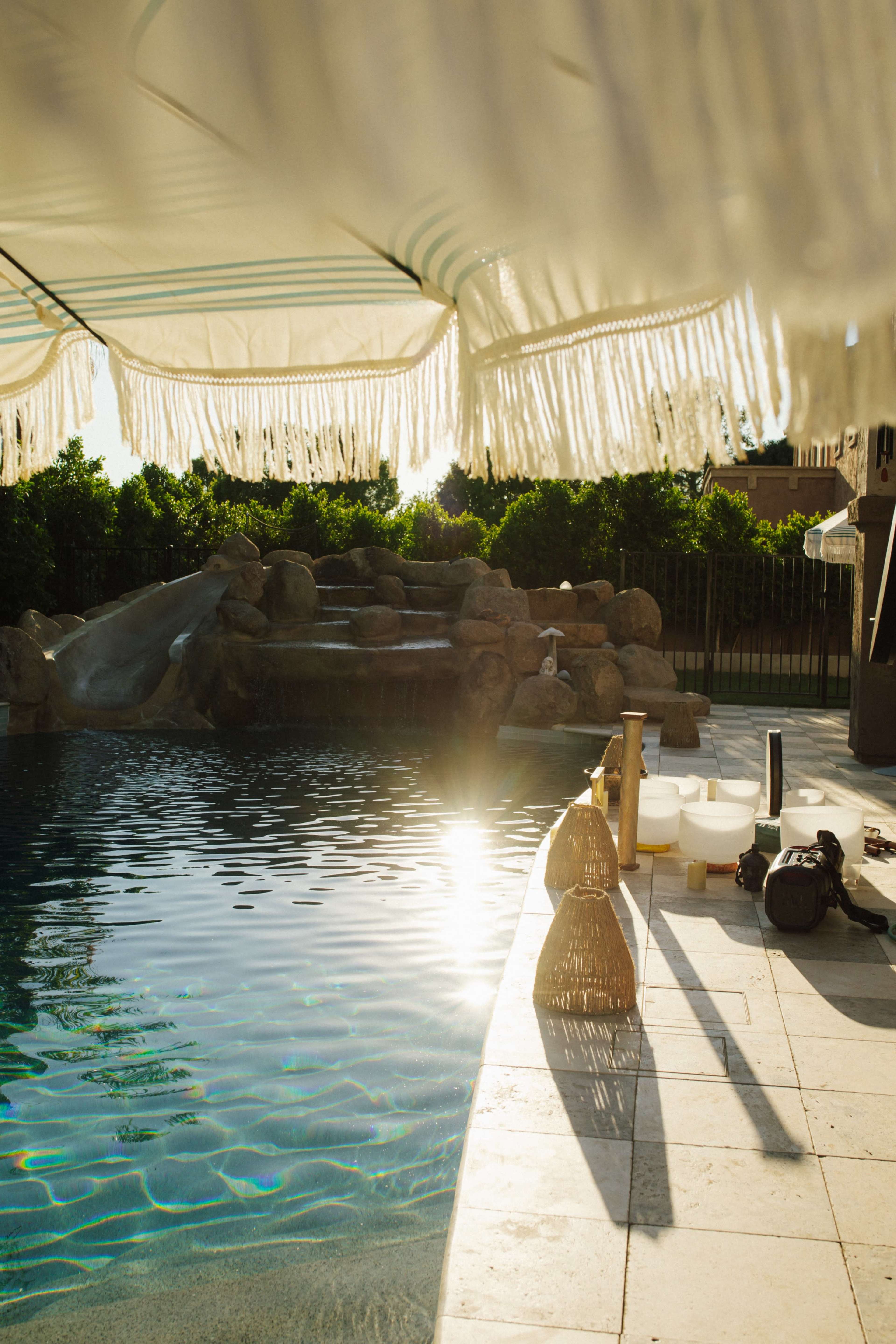 The image shows a swimming pool area with rock features, a shaded canopy, and a reflection of sunlight on the water's surface.